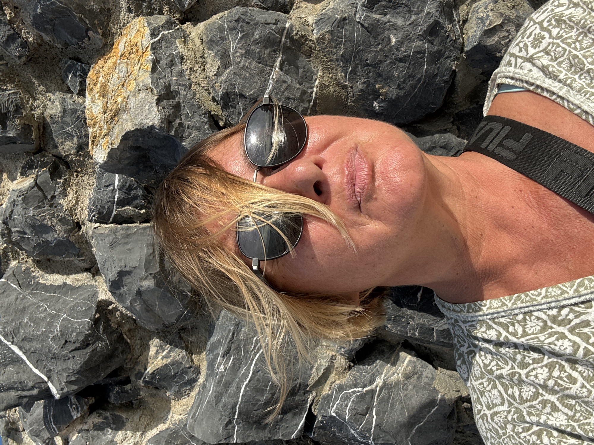 A woman is lying on dark gray rocks with white veins, possibly at a coastal location in Baan Bo Phut, Thailand. She is wearing sunglasses, which partially cover her face, and a patterned top with a black strap visible. The rocks appear to be part of a rocky shoreline or embankment.