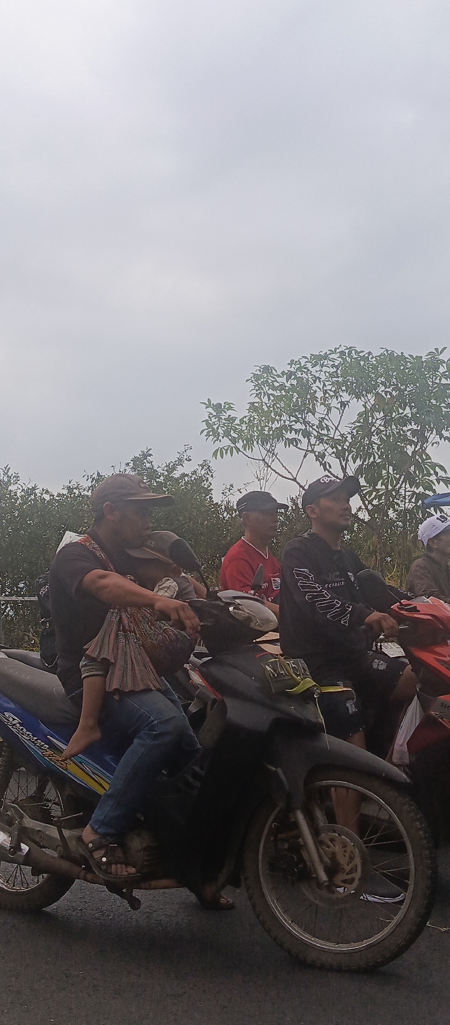 The image captures a group of people on motorcycles in what appears to be a rural or suburban area of Sumbersekar, Indonesia. The sky is overcast, suggesting a cloudy or hazy day. In the foreground, a man wearing a dark t-shirt and a baseball cap is riding a blue and white motorcycle. He is holding a young child, who is wearing a patterned skirt and blue jeans, on his lap. The child's feet are bare. The man is looking down towards the child. Beside them, other individuals are also on motorcycles, facing forward, suggesting they are part of a procession or group traveling together. One man in the middle is wearing a red shirt, and another to his right is wearing a black long-sleeved shirt with white lettering. The motorcycles are parked on an asphalt road, and the background consists of dense green foliage and trees. The overall scene depicts a common mode of transportation and community gathering in the depicted Indonesian locale.