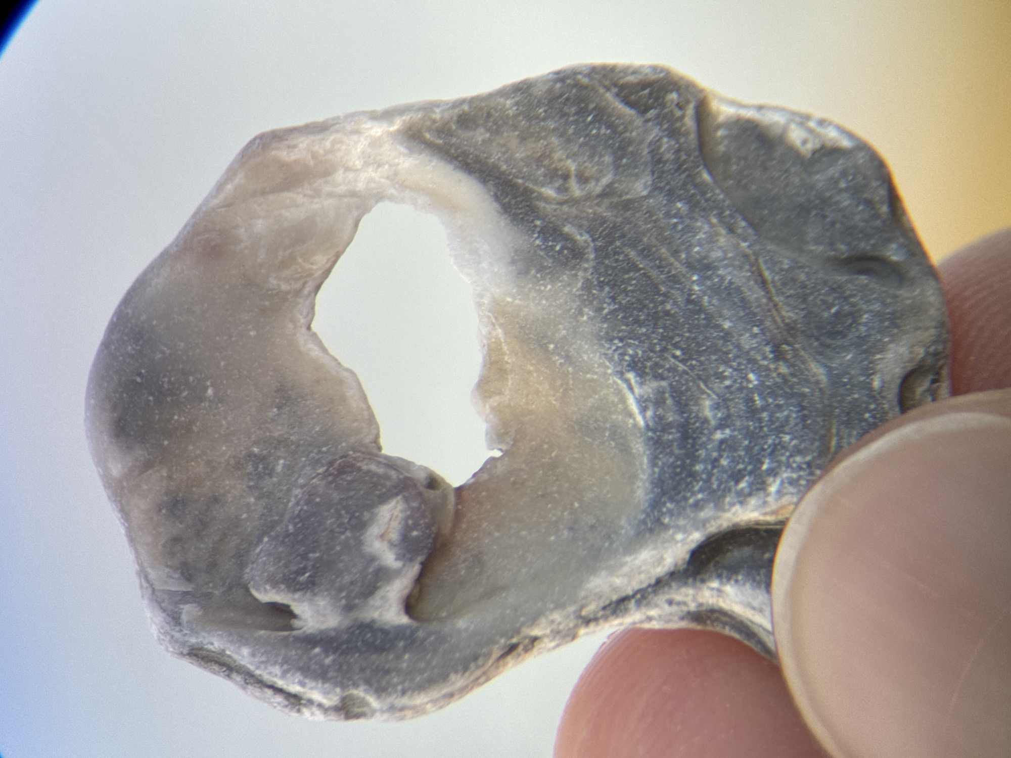 The image is a close-up, macro photograph of a small, irregularly shaped piece of shell with a hole in the center. The shell fragment is held between a thumb and index finger, which are blurred by the close focus. The shell has a greyish-blue exterior with lighter, almost white, striations and a creamy-white interior visible around the hole. The background is a soft, out-of-focus blur of light blue and yellow, suggesting it might be outdoors with a bright sky and sandy ground. There is no indication of Nha Trang, Vietnam, or any urban environment in the image.