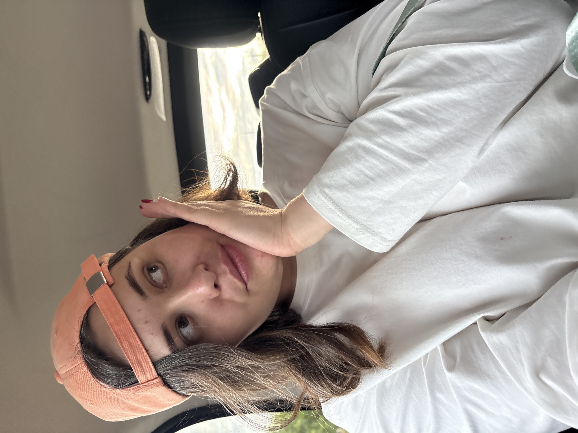 A young woman wearing an orange baseball cap and a white long-sleeved shirt is seated inside a vehicle. She is resting her right hand on her cheek and looking away from the camera. The background shows the interior of a car, with a black headrest visible to the right and a cloudy sky through the window. The lighting suggests it is daytime. There are no identifiable landmarks or indications of the specific location within Zverosovkhoz, Russia.