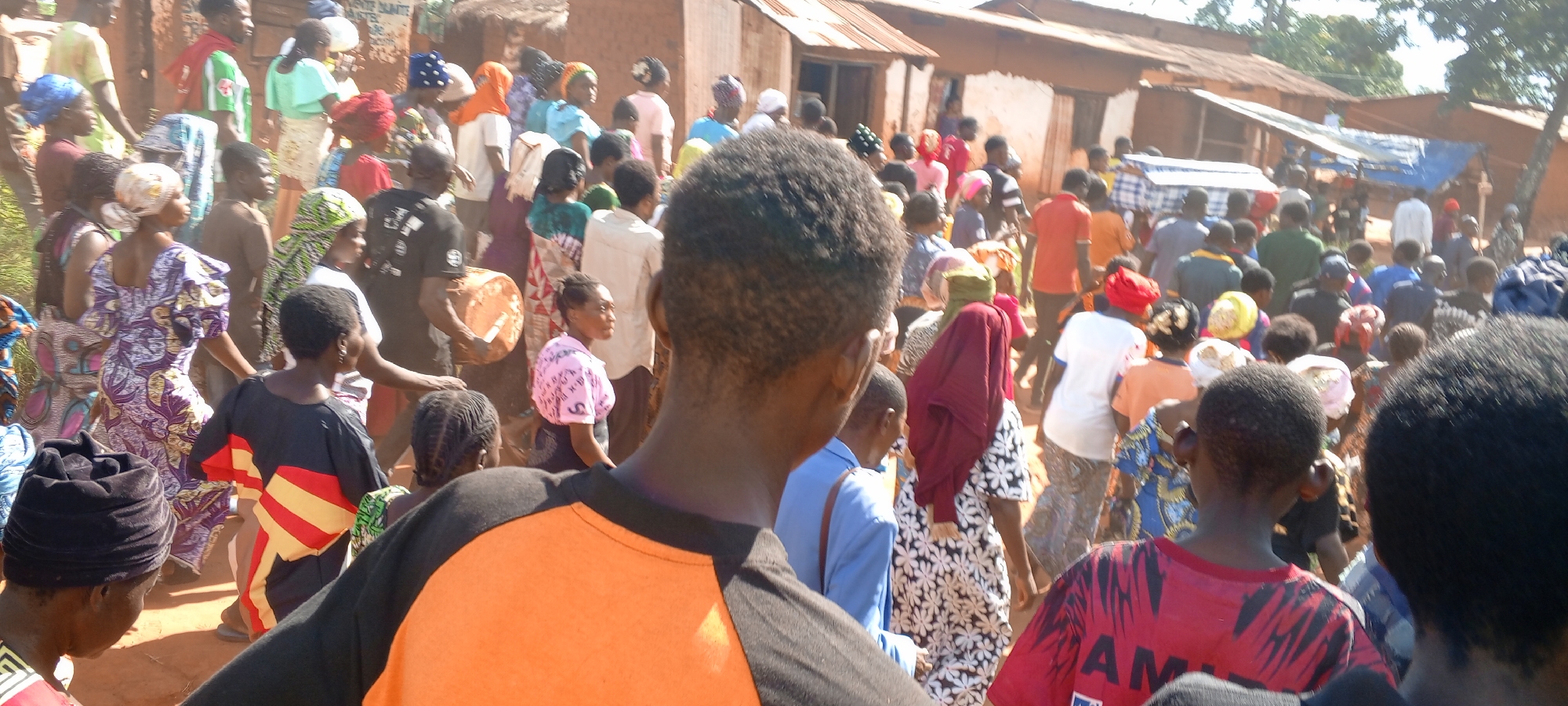 A crowd of people is gathered in Ngalula, Democratic Republic of the Congo. The individuals are diverse in age, gender, and dress, with many wearing colorful traditional clothing and head coverings. The gathering takes place in a rural or semi-rural setting, indicated by simple, low-built structures with corrugated metal roofs in the background. The ground is reddish-brown, suggesting dirt or clay. The mood is indistinct, as faces are obscured, but the density of the crowd implies a community event or gathering.