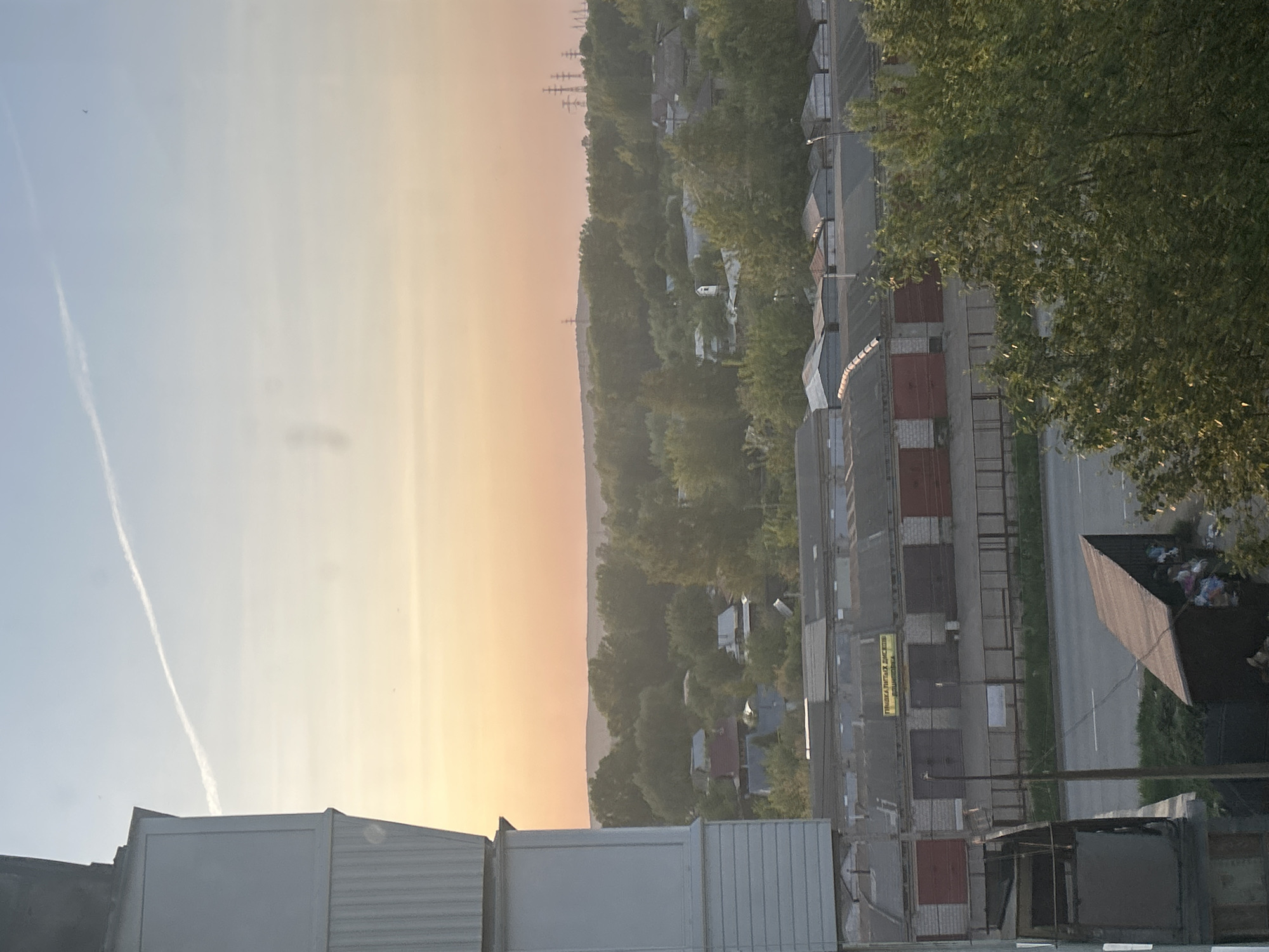 This image shows an elevated view of a suburban area, likely taken from a high building or apartment in Chaikovsky, Russia. The scene is dominated by a long row of brick and metal garages that line a two-lane road. Behind the garages, a dense green area suggests a forest or park, with houses scattered throughout. In the far distance, power lines are visible on the horizon. The sky shows a gradient from light blue to orange and yellow, indicating sunrise or sunset. There is a vapor trail from an airplane. In the foreground are some metal structures, possibly air conditioning units. There are no people visible in the image.