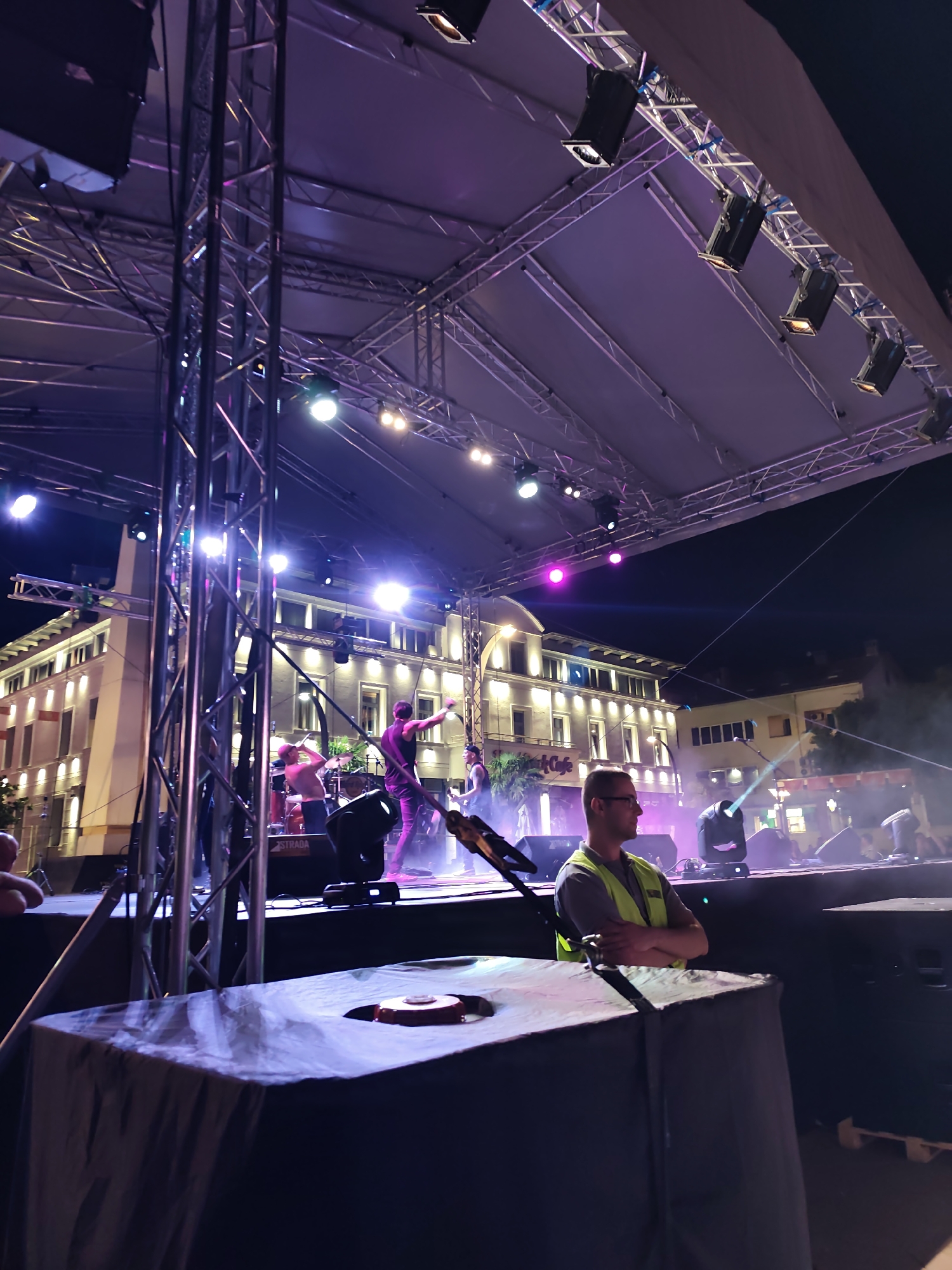 An outdoor night concert or performance is captured from a slightly elevated vantage point, centered on a brightly lit stage in what appears to be a public square in Podgorica, Montenegro. The stage, covered by a light-colored canopy appearing purple from the lighting, is constructed with a metallic truss system supporting numerous stage lights. These lights emit strong beams of white, pink, purple, and blue-green, creating atmospheric haze on stage.
Several musicians are actively performing on stage. To the center-left, a figure in a light top is partially obscured. To the center-right, a musician in a dark tank top is energetically engaged, with one arm raised. A drum kit is visible towards the rear of the stage. Stage monitors and other equipment, including one marked "STRADA," are present on the stage floor.
In the right-center foreground, an adult male wearing glasses and a high-visibility yellow-green vest stands with his arms crossed, facing left towards the stage, observing the performance. He has short hair. Immediately in front of him, a dark-draped table or structure occupies the bottom of the frame, with a circular red, textured object resting on its surface.
The background features a prominent, multi-story building with a light-colored facade and numerous rectangular windows, illuminated by warm, yellow architectural lighting. To the far left, a distinct column-like structure is part of an adjacent building. The sky is dark, indicating the nighttime setting of the event.