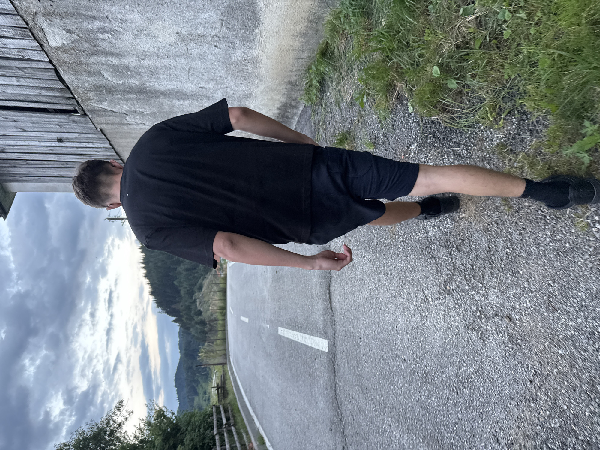 A young man with short brown hair is walking away from the camera on a paved road. He is wearing a black t-shirt, black shorts, black socks, and black shoes. The road has white lane markings. To his right is a rough concrete wall and a weathered wooden structure. To his left is a grassy embankment and a wooden fence, beyond which are trees and hills under a cloudy sky. The image appears to be taken in a rural or semi-rural setting. Based on the context provided, the location is the city of Barutin, Bulgaria.