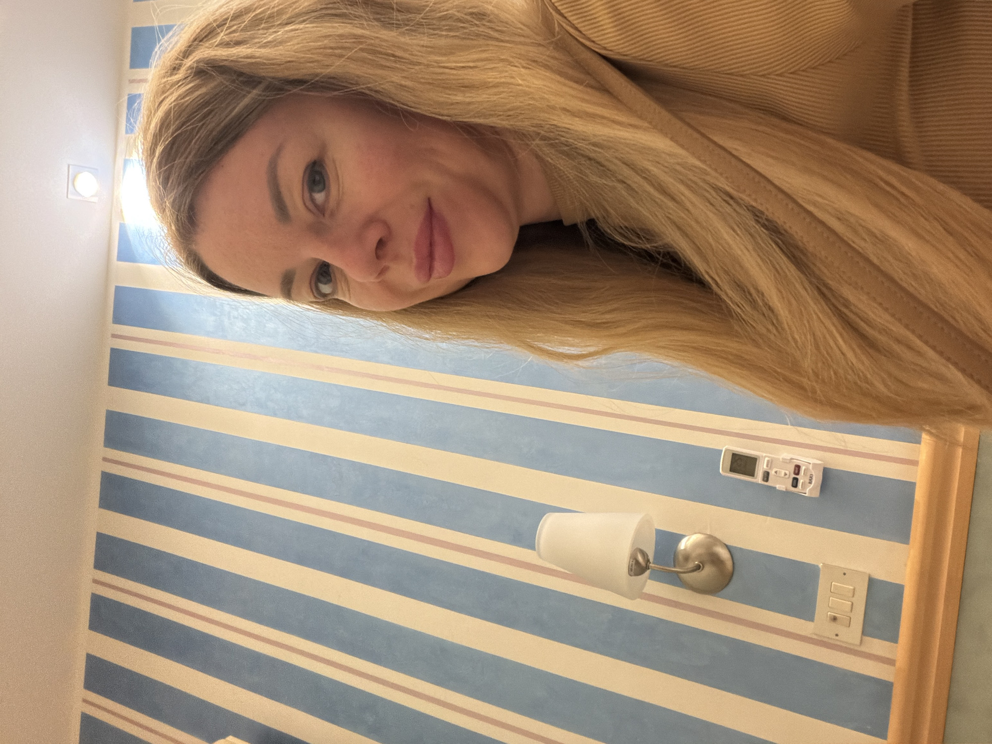 The image shows a medium shot of a blonde woman with fair skin and blue eyes, taken from the chest up. She is looking at the camera and smiling slightly. She has long, wavy blonde hair that is falling over her right shoulder. She is wearing a tan, ribbed turtleneck sweater. The background is a wall with diagonal blue and cream stripes, with thin pink stripes interspersed. To the left of the woman, there is a white lampshade with a silver fixture mounted on the wall. Below the lamp, there is a white electrical outlet with three switches. To the right of the outlet, there is a small white remote control with a screen and buttons, labeled "TAXI." The setting appears to be indoors, possibly a hotel room, in the city of Gatteo, Italy.