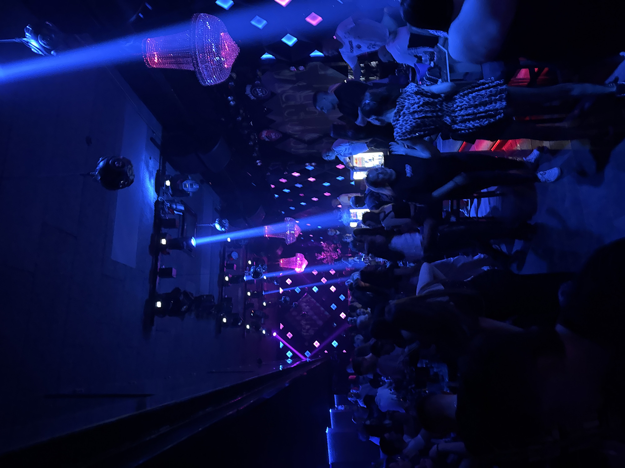 The image depicts the interior of a crowded nightclub. The lighting is predominantly blue, with spotlights and a disco ball contributing to the atmosphere. A chandelier is visible hanging from the ceiling. Many people are present, either standing or seated. Based on the location, the club is likely in Varna, Bulgaria, although no specific evidence is visible.