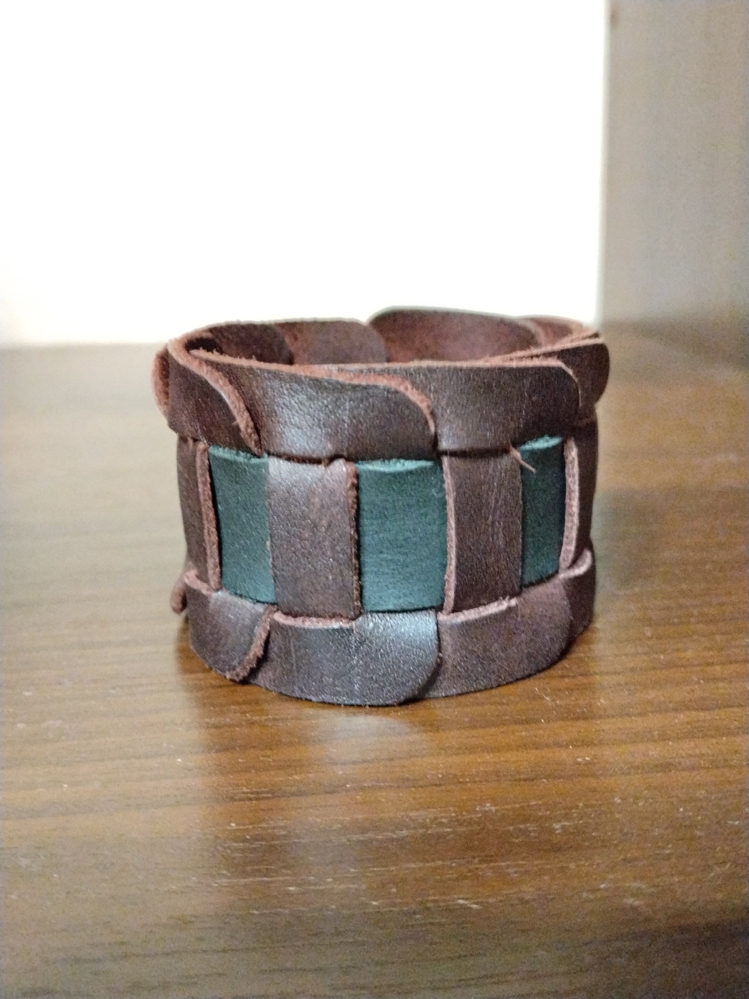 A close-up, eye-level, studio shot shows a brown and green woven leather bracelet resting on a wooden surface. The bracelet is oriented horizontally and its texture is emphasized by the lighting. The background is blurred, with a white area to the left and a lighter gray area to the right, suggesting an indoor setting with a simple backdrop. No people are present in the image. The image does not provide any information about the city of Krasnogorsk, Russia.