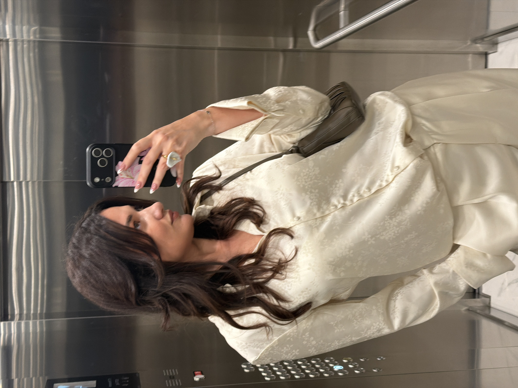 A woman with long, dark brown wavy hair is taking a selfie with her phone in an elevator. She is wearing a light yellow patterned button-up shirt or blouse and is holding a black smartphone with a floral design on the case. She has a ring on her finger with a white flower-shaped design. The elevator has a stainless steel interior with buttons visible on the lower part of the wall. The environment is simple and enclosed, focusing attention on the woman and her action. The location cannot be determined solely from the visual elements in the photo; no elements indicate the city of Belgrade, Serbia.