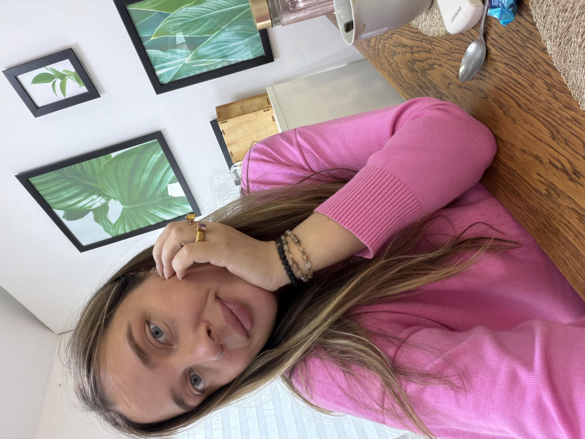 A young woman with long brown hair and blue eyes, wearing a pink sweater, is shown in an indoor setting. She is wearing rings and bracelets. Behind her on the wall hang three framed pictures of green leaves. A wooden table is partially visible, on which is a spoon, a small container labeled "Lesser," a food item in a blue wrapper, and a part of a blender. The background is a white wall and a white vertical blind. The image does not provide information about the city of Chaikovsky, Russia.