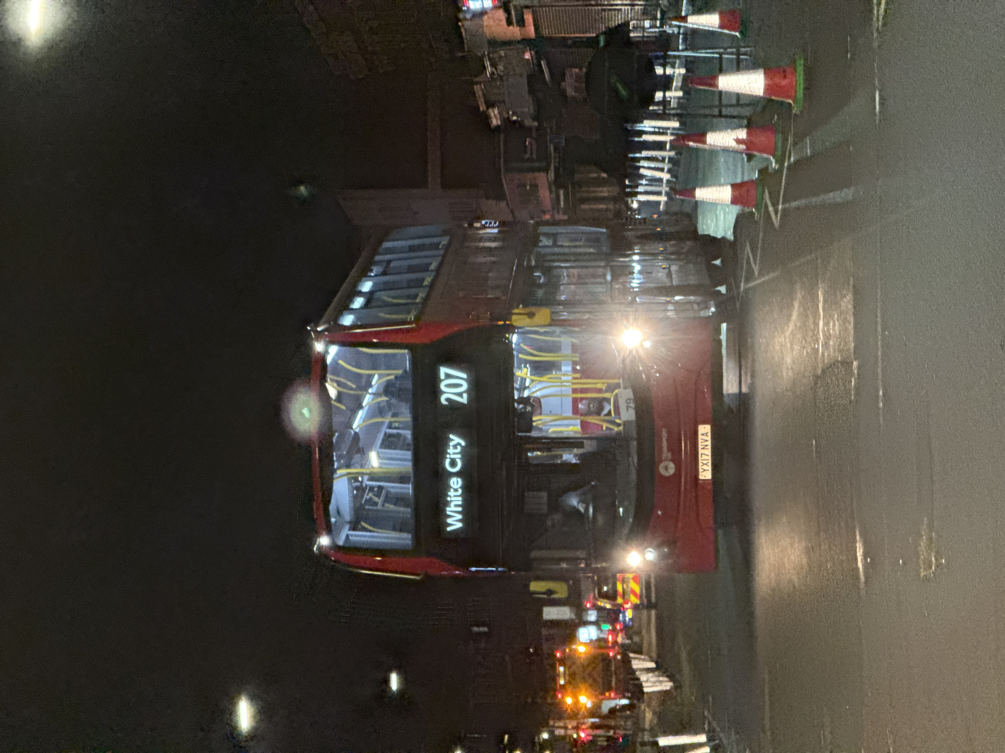 The image depicts a red double-decker bus at night in London, United Kingdom. The bus is a "White City" route 207, with the license plate "YX17 NYA". The road is wet and reflects light. Red and white striped traffic cones are visible on the right side of the frame. There is an ambulance parked behind the bus with lights flashing. Streetlights are on, but the overall environment is dark. The image is taken at an unusual angle, possibly from a low vantage point.