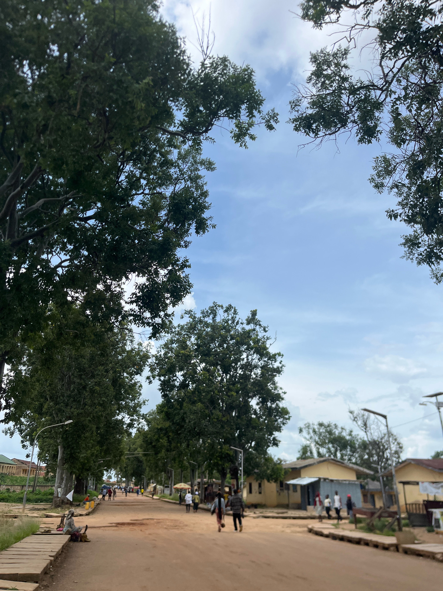 An eye-level, medium shot captures a dusty, unpaved road in Rafin Par, Nigeria, framed by lush green trees on either side and a partly cloudy sky overhead. The road, appearing to be a main thoroughfare, is lined with modest, single-story buildings with yellow facades and corrugated metal roofs. Several individuals are present, walking along the road or gathered near structures. On the left side of the road, a woman is seated on a wooden plank pathway, with a basket beside her. Further down the road, a few people are walking in both directions, and a person is seated on a bench near a tree. The scene suggests a typical day in a Nigerian town, with daily life unfolding along the street.