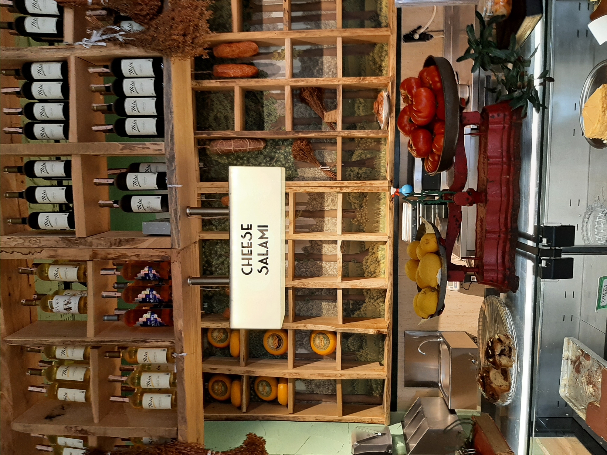The image depicts the interior of a food establishment, likely a deli or market, characterized by wooden shelves displaying various food items and drinks. On the upper shelves, numerous bottles, predominantly wine bottles with white and black labels, are neatly arranged. Several bottles appear to be white wine, while others are dark, possibly red wine. Below these, on more shelves, are round cheeses with yellow rinds and labeling. In the center of the image, a brightly lit sign reads "CHEESE SALAMI." To the right of the sign, hanging from hooks, are several cured meats, likely salami.
The lower part of the image shows a counter area. A vintage red scale is visible, with a pile of lemons on the left side and a pile of tomatoes on the right. A glass platter containing sliced baked goods, possibly cake or bread, is positioned in front of the scale. Further to the left, a stainless steel sink with a faucet is mounted into the counter. In the foreground, a metal tray with some remaining food residue is visible, and to the far right, a book and a glass jar containing preserves can be seen. The overall environment is rustic, with wooden elements and a backdrop that appears to be a printed image of a forest. There are no people visible in the frame. The location is identified as Alanya, Turkey.