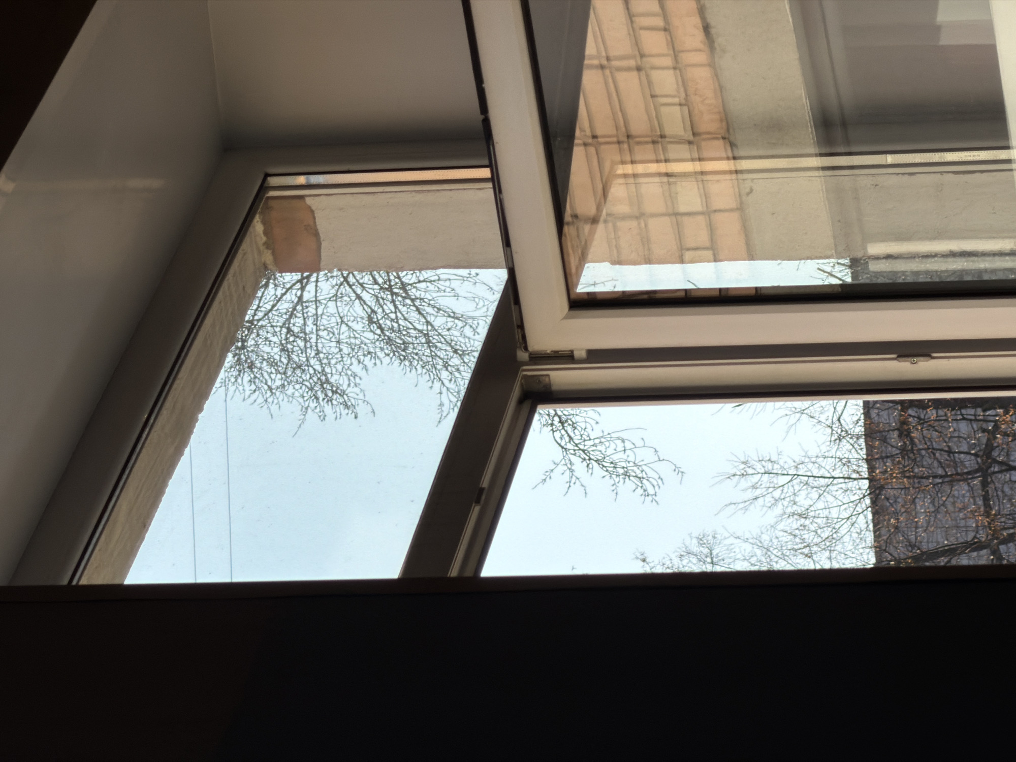 The image shows an open window looking out at an overcast sky and bare tree branches. On the right, a building is visible through the window. The window frame is visible, as is part of the wall surrounding the window. The dark bottom portion of the image appears to be a countertop or ledge. It is difficult to definitively confirm the location is Izhevsk, Russia without more context or visual cues specific to that city.