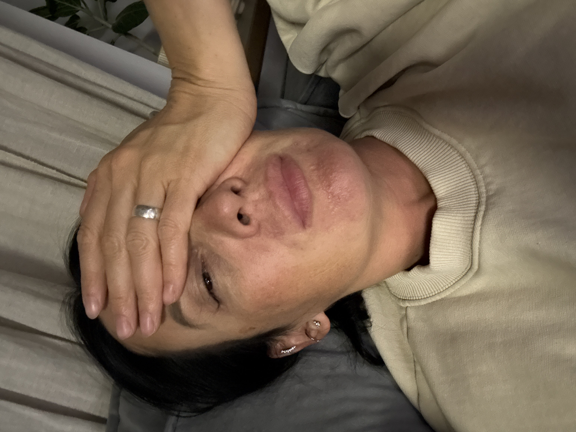 A woman is lying down, facing the camera with her hand covering part of her face. She wears a ring on her finger and earrings. She is wearing a tan-colored sweatshirt and resting on gray bedding. A plant can be seen in the background. There is no verifiable information suggesting a location of Chaikovsky, Russia.