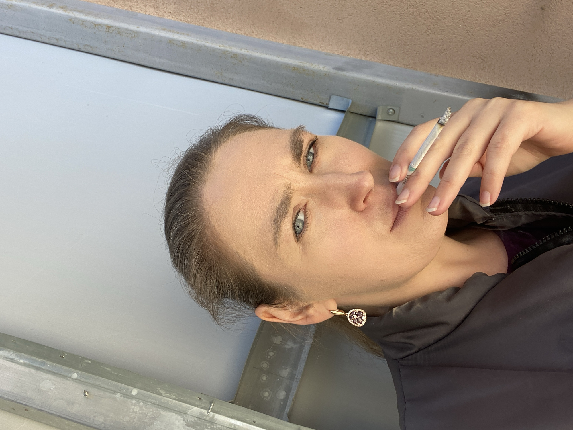A close-up, tilted portrait shows a woman holding a lit cigarette. She has fair skin, blue-green eyes, and light brown hair with visible grey strands, pulled back from her face. Her lips are slightly parted, and she gazes directly towards the viewer with a neutral expression. She wears a dark, possibly padded, jacket and a purple garment underneath. A gold-toned hoop earring with a cluster of dark red/purple stones surrounded by tiny clear gems is visible in her right ear.
She holds the cigarette between the index and middle fingers of her right hand. The cigarette has a white filter with green text, which appears to read "Vogue." Ash has accumulated on the tip of the cigarette.
The background consists of a light grey, plain wall or surface, topped by a horizontal metallic beam or railing. Above the beam, a light brown, textured surface is visible. The scene suggests an outdoor or semi-outdoor location, possibly against a building or structure in Yekaterinburg, Russia. The lighting is natural, indicating daylight, and her jacket suggests cooler weather.