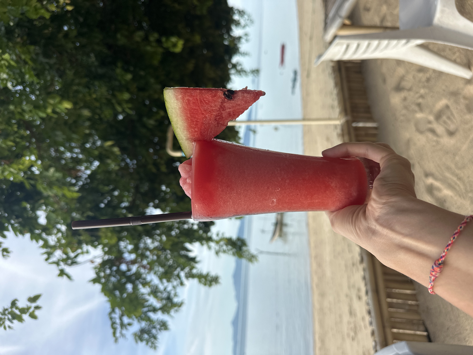 A person's hand holds a tall, clear glass filled with a vibrant red, icy beverage, likely a watermelon smoothie. A dark brown straw extends from the drink. A triangular slice of watermelon, showing its pink flesh and green rind, is perched on the rim of the glass. The background depicts a serene beach scene in Baan Thong Po, Thailand. The sand is light brown, with footprints visible. Beyond the sand, the calm blue ocean stretches towards a distant, hazy shoreline with faint mountains. A small boat floats on the water. Lush green trees with dappled sunlight frame the top portion of the image, creating a shaded canopy. The overall atmosphere suggests a relaxed, tropical vacation setting.