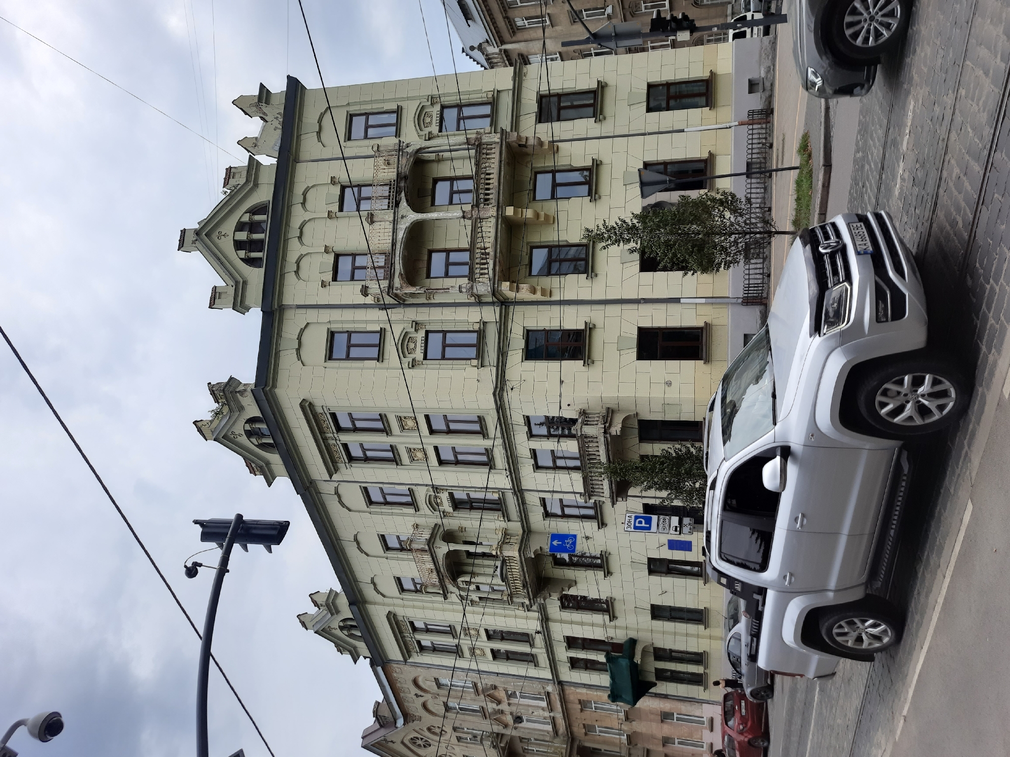 The image captures a street scene in Lviv, Ukraine, with a prominent pale yellow building dominating the frame. This building, featuring multiple stories with ornate balconies and arched window designs, suggests an architectural style common in historical European cities. In the foreground, a silver pickup truck is driving on a cobblestone street. Several other vehicles, including a red car and a grey sedan, are parked or moving on the road. Overhead, traffic lights and utility wires crisscross the cloudy sky. Signage, including a blue sign with a bicycle icon and a parking sign with Ukrainian text, indicates urban infrastructure. The overall atmosphere is that of a typical day in a European city with a blend of old and new elements.