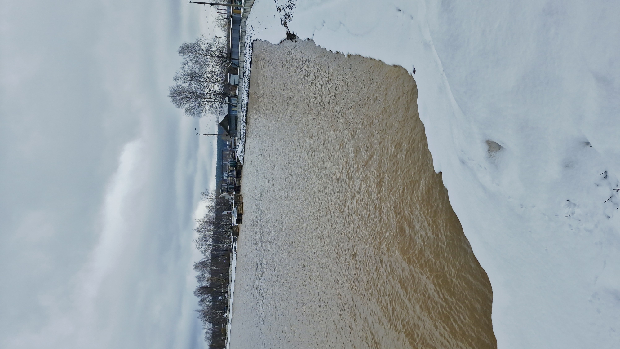 The image depicts a winter river scene in Otchor, Russia. A wide river, brown in color, flows vertically through the frame, with snow-covered banks on either side. The left bank has trees lining the edge, and behind the trees are buildings and lampposts suggesting a town or urban area. The sky is overcast. The river appears to be swollen. There is no sign of people or animals.