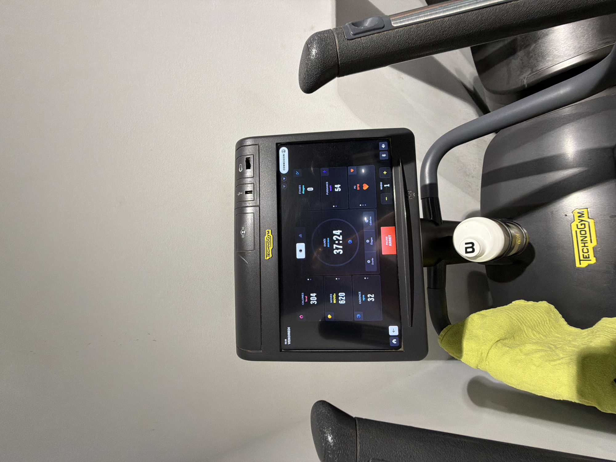 This is a close-up, slightly angled shot of a modern stationary exercise bike's console, indoors. The bike is black with grey handlebars and a prominent touchscreen display. The screen shows workout metrics, including time elapsed (37:24), calories burned (304 kcal), moves (620), and cadence (32 spm). It also indicates the current resistance level (NIVEAU 1) and heart rate (FC BPM), though the heart rate is not specified. The word "TECHNOGYM" is visible on the console and the side of the machine, identifying the brand. A bright yellow cloth is draped over a part of the bike's frame near the screen. The background is a plain, light-colored wall, suggesting an indoor gym or fitness area. The time displayed on the screen is 15:18, and the day is "VENDREDI" (Friday). There are no people visible in the image. The lighting is even, indicating artificial light.