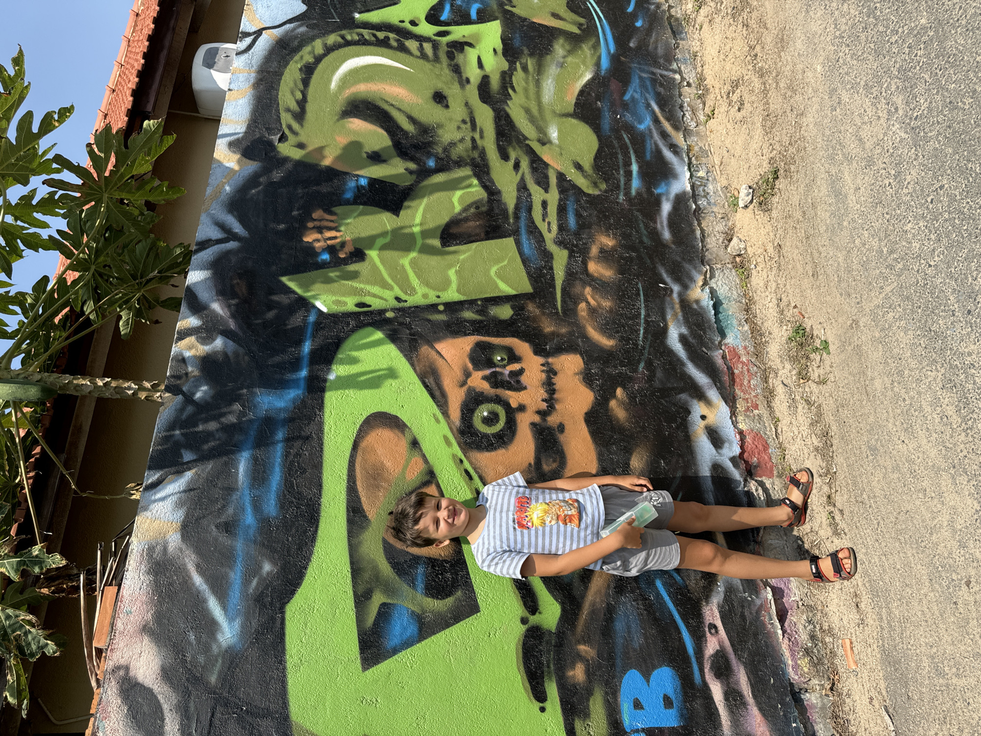 A young boy is lying against a wall covered in graffiti in Mũi Né, Vietnam. He is wearing a striped t-shirt with a cartoon character on it, grey shorts, and sandals. The graffiti on the wall is a mix of green, black, blue, and brown, depicting abstract shapes and what appears to be a skull. Part of a building is visible at the top left, including the roof and some foliage. To the right of the wall is a paved road. The sky is visible in the upper left corner. The boy is holding a light-colored rectangular object in his right hand. He is smiling.