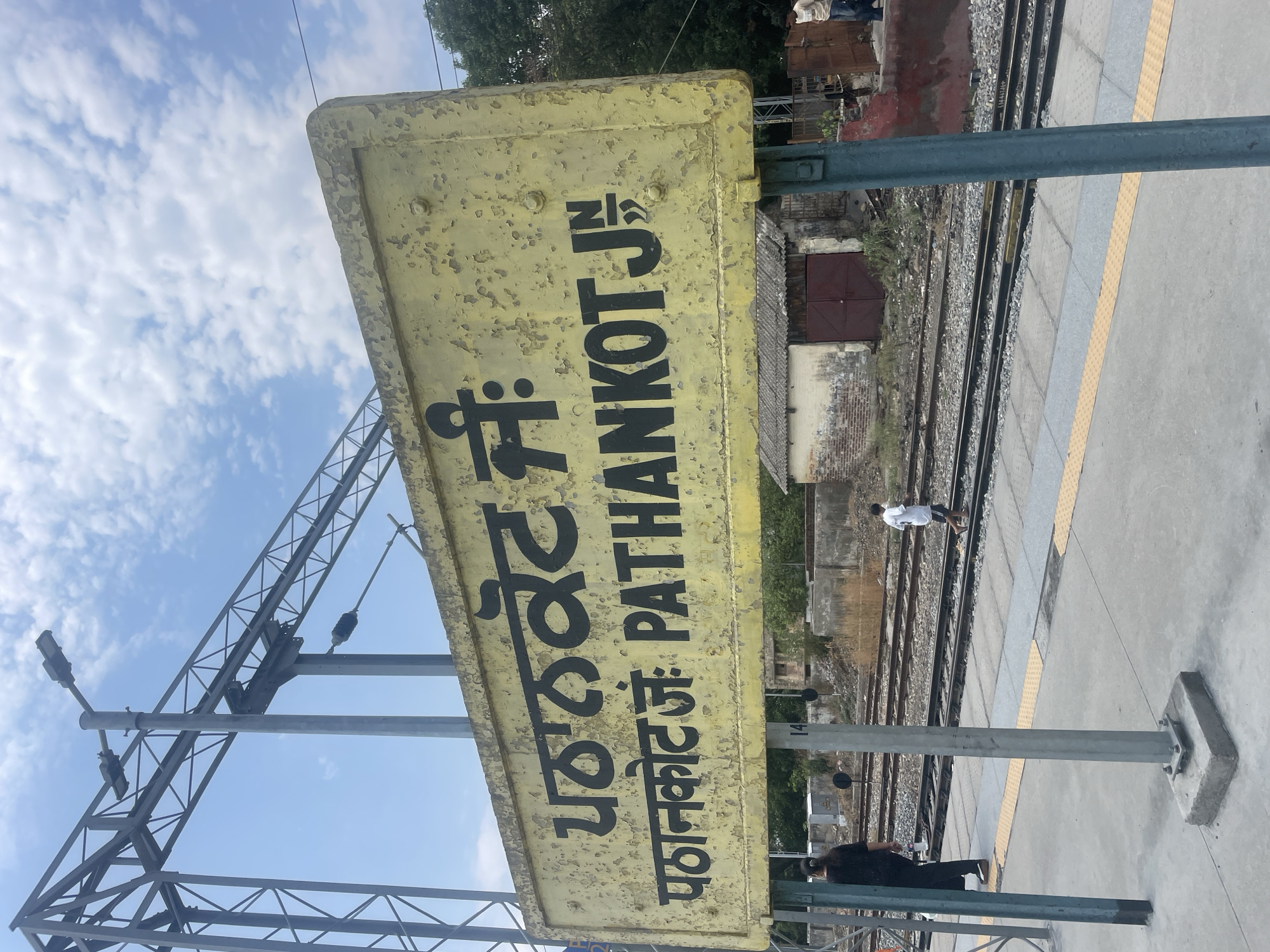 The image shows a weathered yellow sign at the Pathankot Junction railway station in Pathankot, India. The sign displays the station name in Punjabi script and English. Several railway tracks are visible behind the sign, with a platform in the foreground. A few people are present on the platform and near the tracks. The sky is cloudy.