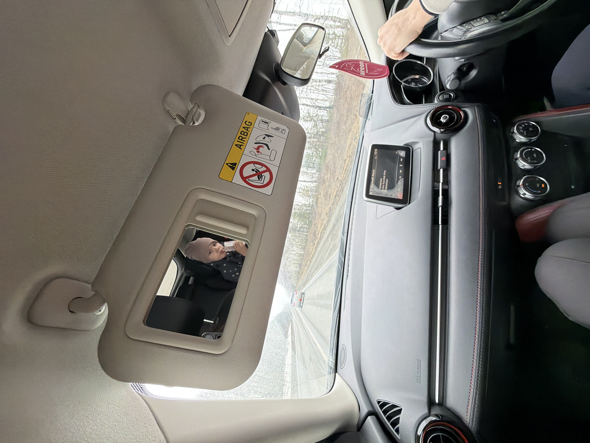 A view from inside a car shows the driver's side. The visor is flipped down and its mirror reflects a baby in a car seat, holding a bottle to their mouth. An air freshener hangs from the rearview mirror. The car's dashboard is visible, including the steering wheel, infotainment screen, and climate control knobs. The location is indeterminable.