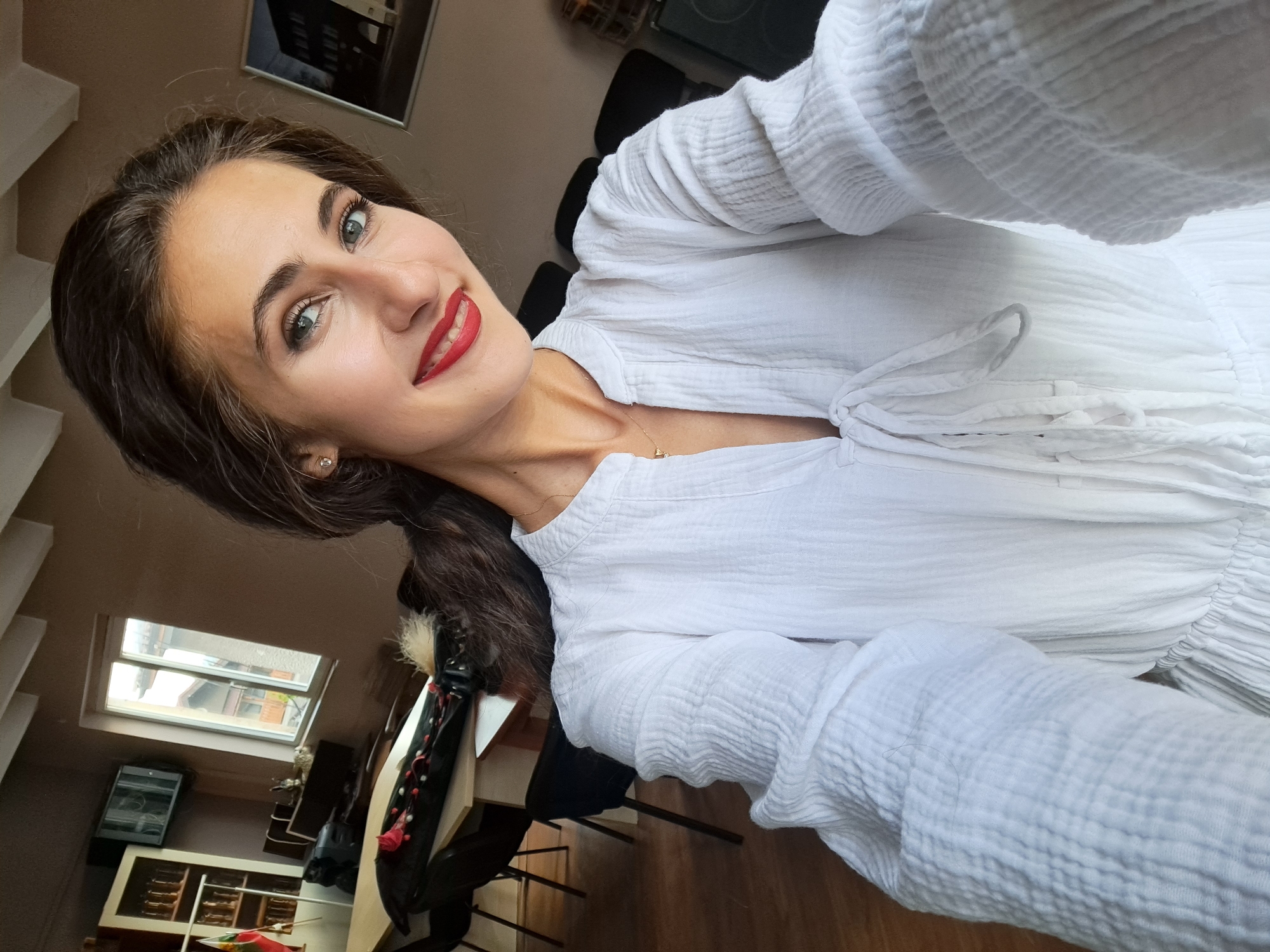 The image is a frontal, eye-level selfie of a young adult female, taken indoors in the city of Voynegovtsi, Bulgaria. The subject has dark hair, styled down, and is wearing red lipstick and visible eye makeup. Her blue-green eyes are directed at the camera, and she displays a wide smile, showing her upper teeth. She is attired in a white, textured garment, possibly crinkled cotton, featuring a V-neckline with decorative ties and long, voluminous sleeves with elasticized cuffs. A thin gold chain necklace with a small pendant is visible, along with a small stud earring in her left ear.
The background reveals an interior space with a warm brown wall color and white ceiling beams. To the subject's left, a wooden table is present with dark chairs. On the table, a dark, elongated decorative object with red and white accents, and a patch of light-colored fur or fluff, is visible. Behind the table, a white shelving unit holds multiple clear glass bottles or jars, and a partially visible flag with green, white, and red stripes is displayed. A window behind this arrangement overlooks rooftops and what appears to be residential buildings. Above the shelving unit, a television monitor is mounted on the wall. To the subject's right, a framed picture depicting a dark building is mounted on the wall. The room is illuminated by natural light.