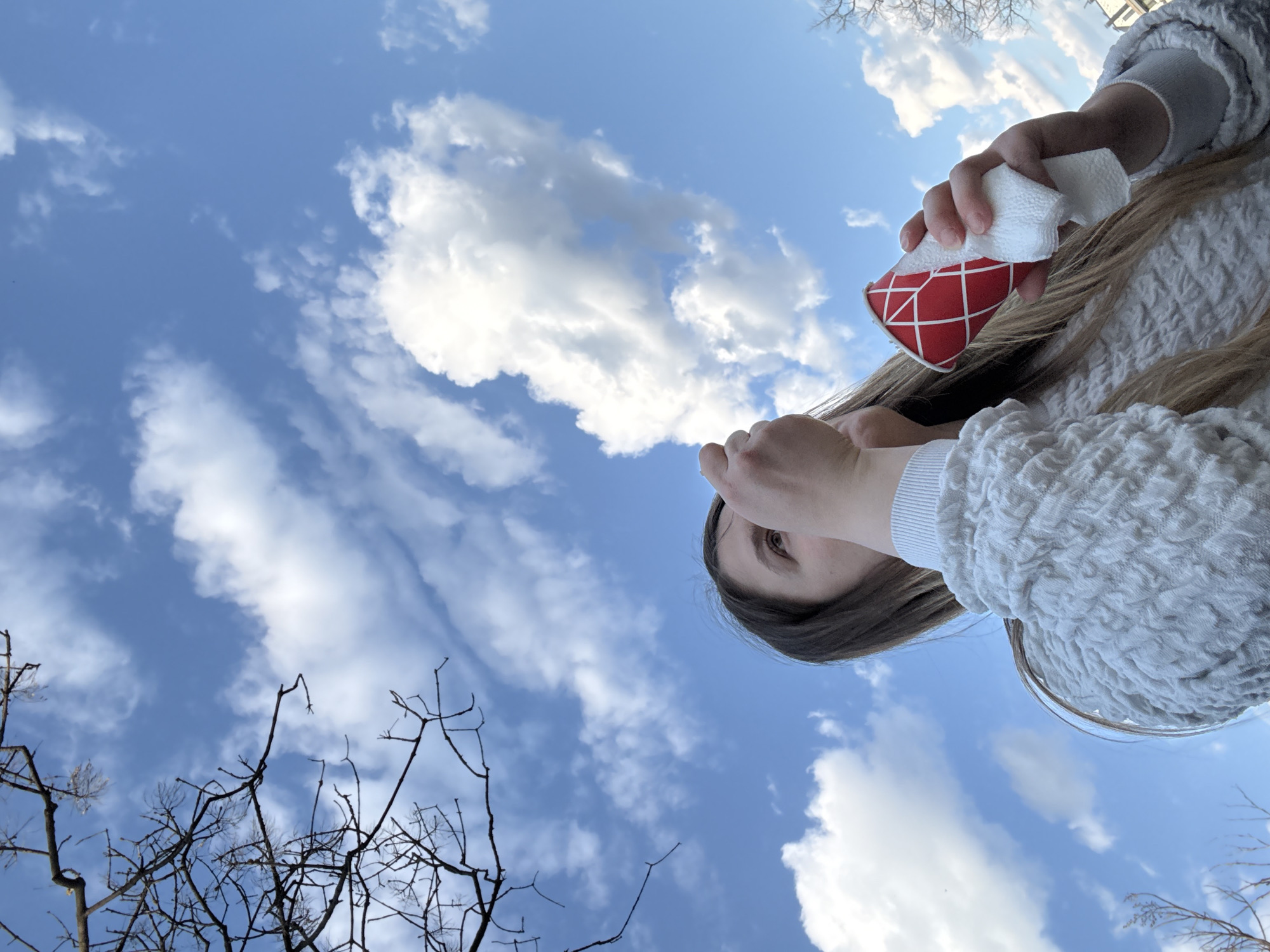 A low-angle shot of a person holding a red and white patterned cup with a white tissue. The person is wearing a white sweater and has brown hair. The background is a bright blue sky with scattered white clouds and bare tree branches in the lower left corner. The location is in Stara Zagora, Bulgaria.