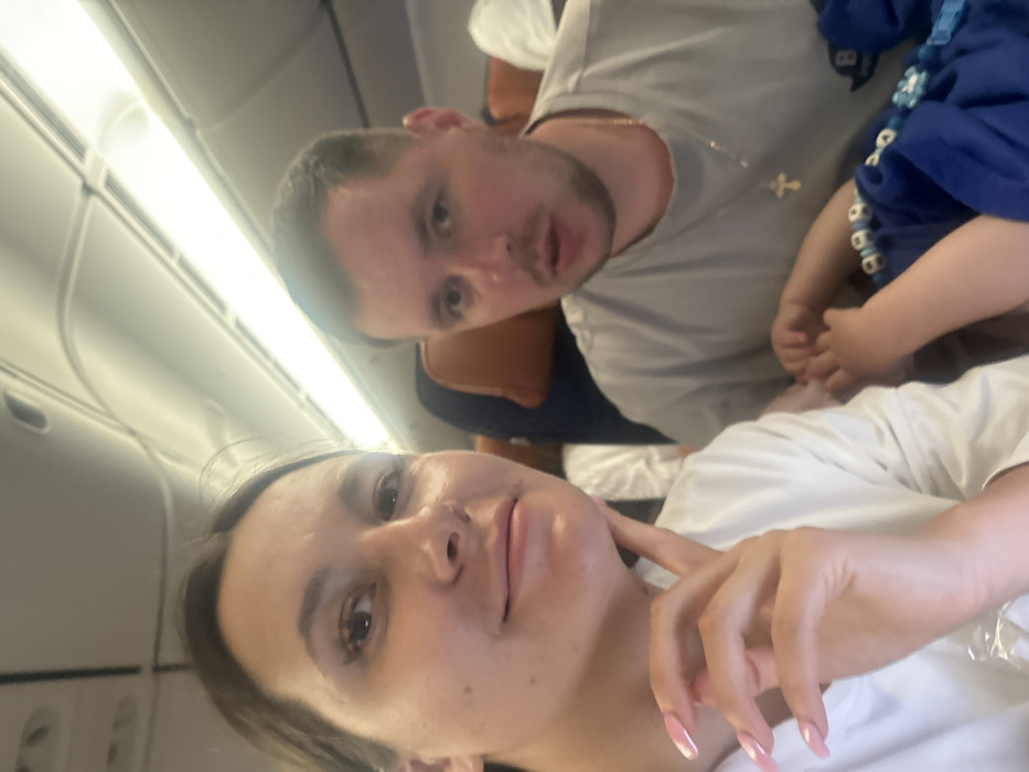 The image captures a selfie-style shot of what appears to be a family inside an airplane cabin. The photograph is oriented vertically, with the subjects appearing rotated 90 degrees clockwise relative to the typical horizontal plane of an aircraft interior.
In the immediate foreground, a woman's face is prominent, taking up the lower half of the rotated frame. She has dark hair, and her eyes are looking directly at the camera. Her expression is neutral to slightly smiling. Her right hand is visible, with manicured nails that are long and have a light pink or white ombré design, resting near her chin. She is wearing a light-colored, possibly white, t-shirt.
Behind and slightly above the woman, a man's face and upper torso are visible. He has short dark hair and is also looking towards the camera, with his mouth slightly ajar as if he might be speaking or just about to. He is wearing a light grey t-shirt and a thin gold chain with a small cross pendant around his neck. His arm and shoulder are visible, suggesting he is seated next to or slightly behind the woman.
To the right of the man, and partially obscuring him, are glimpses of at least one child, possibly two. One child's hands and arms, dressed in a blue top and wearing a blue beaded necklace or accessory, are visible near the man's chest. Another child's head, wearing a white top, is partially seen in the very bottom right corner of the image, close to the woman's side.
The background clearly shows the interior of an airplane. The characteristic curved ceiling with overhead compartments and bright, illuminated light strips running along the length of the cabin are visible. The seats are only partially seen, with brown headrests visible behind the man. The overall lighting is bright, suggesting daytime or well-lit cabin conditions. No text is visible in the image. The scene conveys a casual moment captured during travel, likely a family vacation or trip.