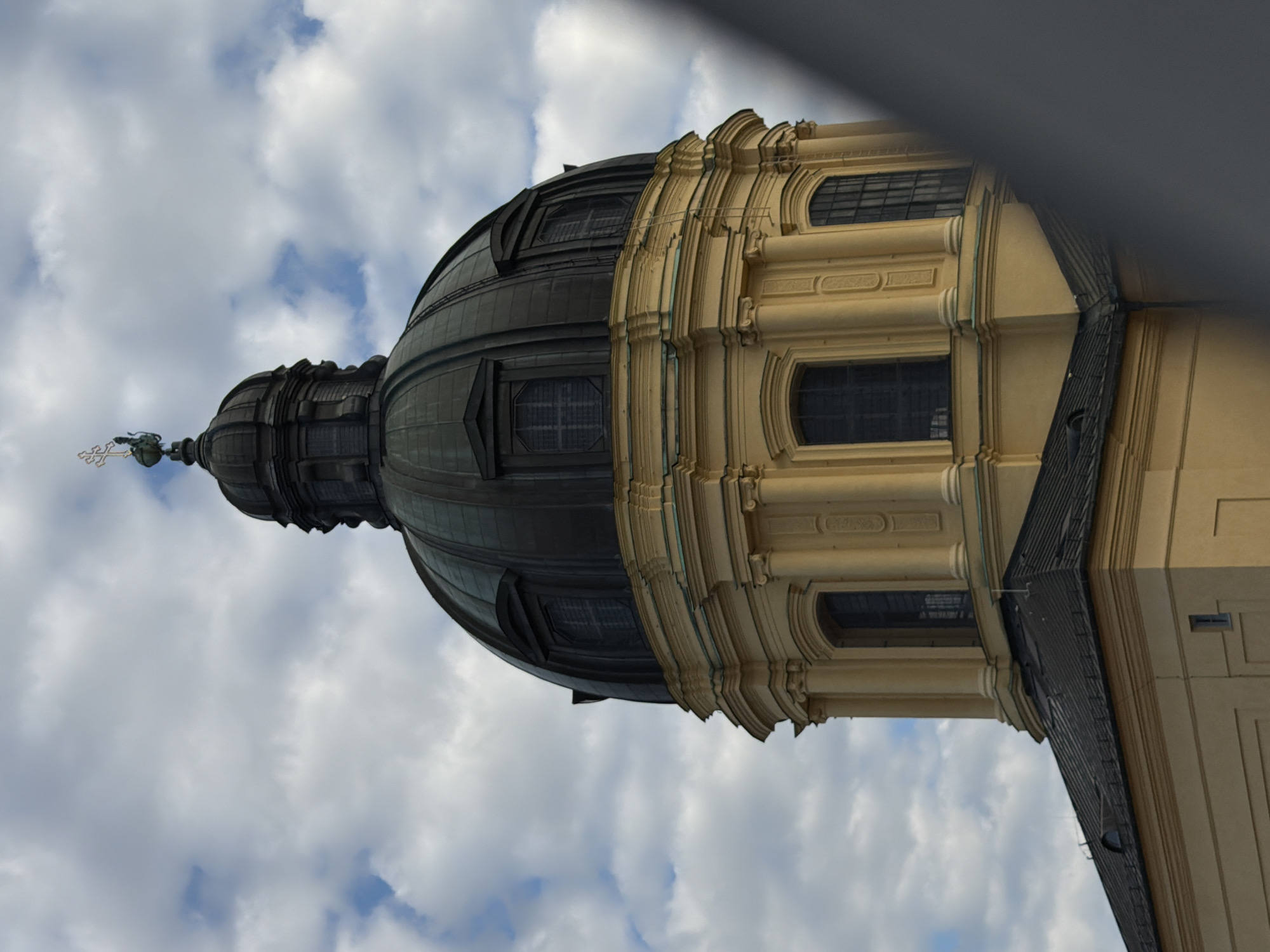 The image shows a close-up, eye-level, vertical shot of the dome of the Theatinerkirche in Munich, Germany. The dome is dark green with a yellow base, and it is topped with a cross. The sky is cloudy with patches of blue visible. The lower part of the image shows the roof of the church, which is also dark green. The Theatinerkirche is a prominent landmark in Munich.