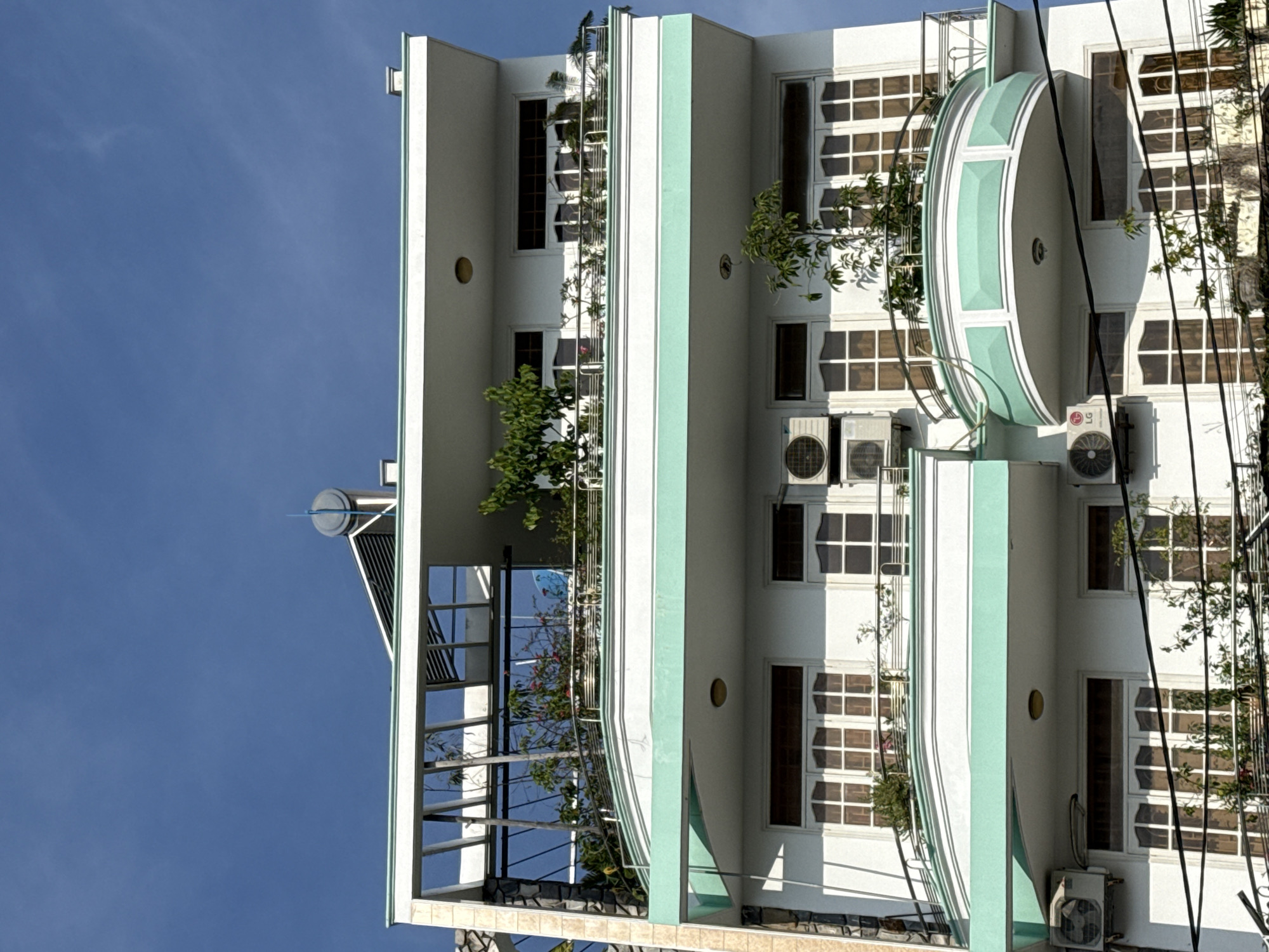 The image depicts a modern, multi-story building in Bac Nha Trang Ward, Vietnam, under a clear blue sky. The building features multiple balconies with white railings and light green trim. Plants are visible on many of the balconies, adding greenery to the structure. Several windows with white frames and multiple panes are present on each level. Air conditioning units are mounted on the exterior walls of the building. The top of the building has a flat roof with some equipment, possibly related to solar water heating. The building appears to be a residential or mixed-use property.