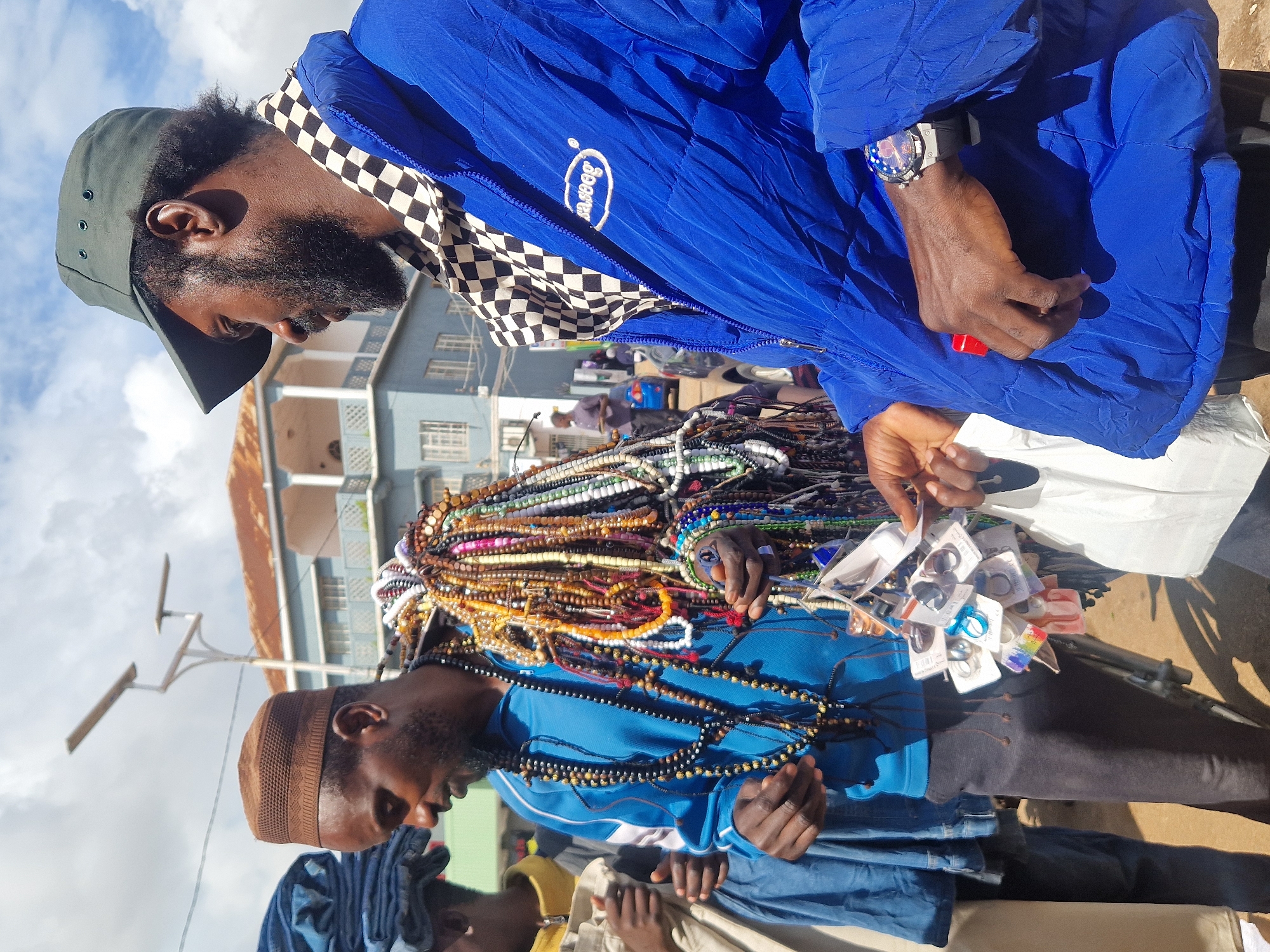Two men are featured in the foreground of the image, likely in a marketplace setting. The man on the left wears a brown kufi and a blue zip-up jacket, and is carrying a large bundle of beaded necklaces and bracelets around his neck and shoulders. He holds a selection of small, packaged items, possibly accessories, in his right hand. The man on the right, wearing a dark green cap and a blue puffer jacket over a checkered shirt, has a beard and a watch on his left wrist. He holds a small red object and a white bag in his hands. In the background, a light blue building with white balconies and windows is visible, along with streetlights and parked cars. The sky is partly cloudy.