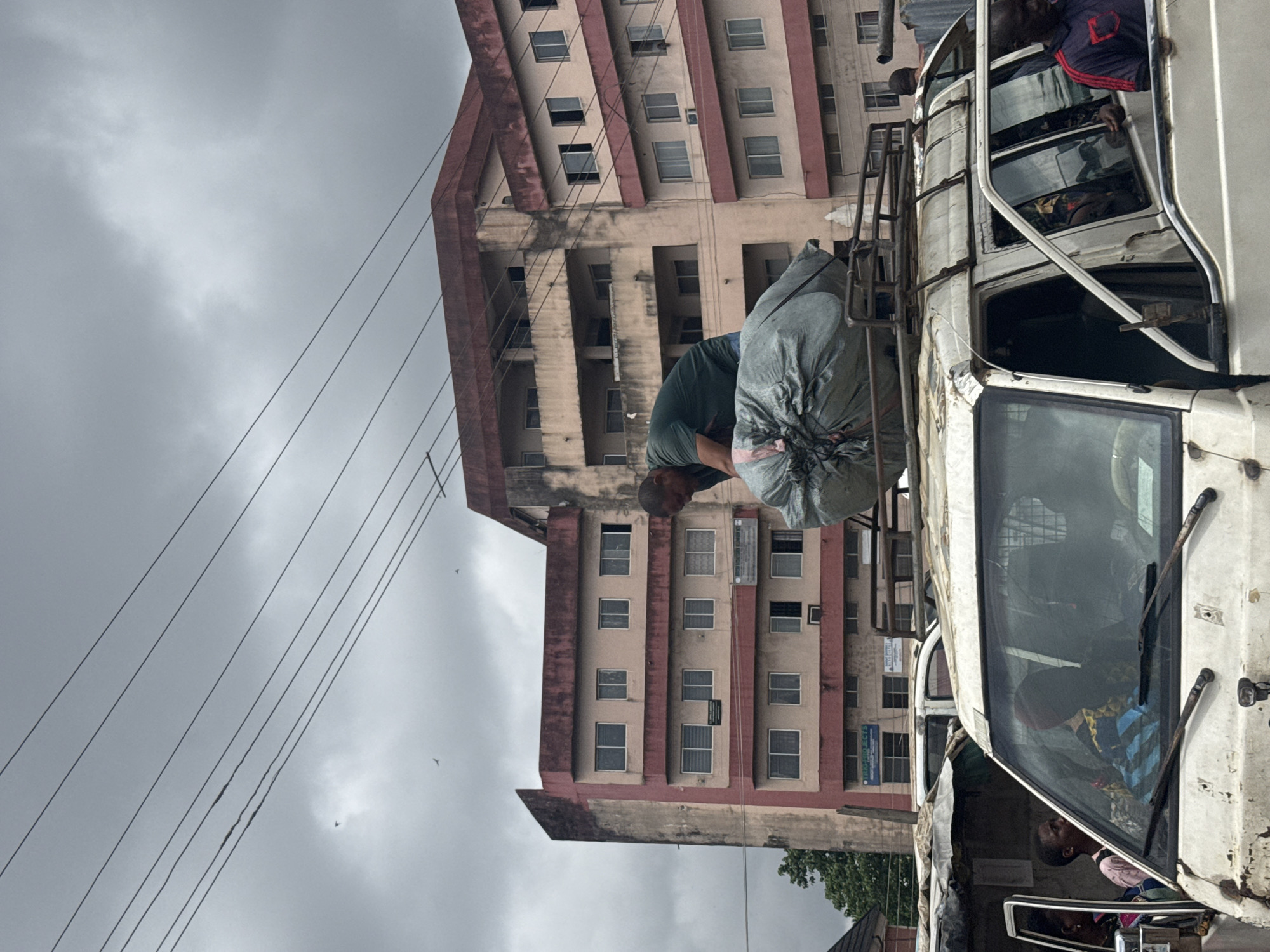 The image captures a cloudy day scene in Owerri, Nigeria, featuring a man on top of a white minivan, securing a large, gray, tarp-wrapped bundle onto the vehicle's roof rack. The minivan is parked in front of a faded, multi-story pink and cream-colored building with numerous windows. Several people are visible inside and around the minivan. One person, wearing a red and blue striped shirt, is standing next to the open passenger door. The sky above is overcast, with dark clouds, and several power lines crisscross the upper portion of the frame. The overall environment appears to be an urban setting, possibly a street or a busy thoroughfare, with a slightly dilapidated feel to the building and the minivan.