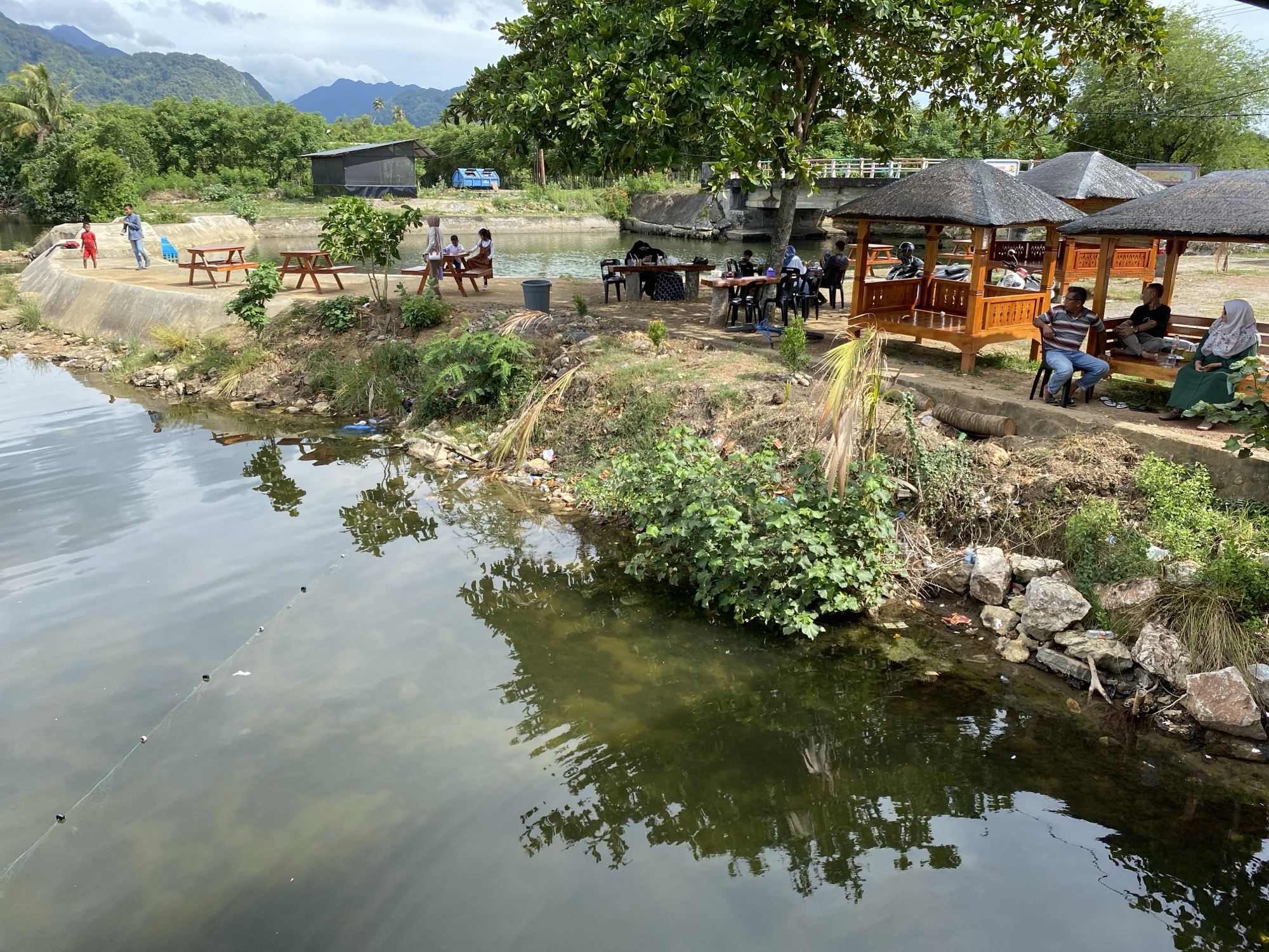 This image captures a waterfront area in Aneuk Paya, Indonesia, featuring a tranquil river or canal bordered by lush greenery and a mountainous backdrop. Several wooden picnic tables and benches are strategically placed along the bank, some occupied by people. To the right, three traditional wooden gazebos with thatched roofs offer shade, with patrons seated at tables within and around them. In the middle ground, a bridge spans the water, and a few structures are visible in the distance. The water reflects the surrounding trees and sky, creating a serene atmosphere. A fishing line with buoys is visible stretched across the water in the foreground. The overall scene depicts a peaceful outdoor recreational space, likely a local gathering spot.