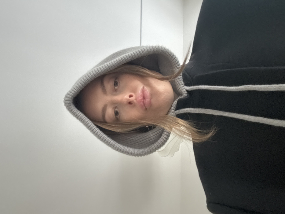 An indoor, eye-level selfie shows a single individual, gender indeterminate, from the upper chest to the top of the head. The individual has light-colored hair, a fair complexion, and is wearing a black garment, possibly a coat or hoodie, featuring a gray ribbed knit hood that is pulled over their head. Two vertical gray drawstrings are visible at the front of the garment. The individual's lips are slightly puckered, and they are looking directly at the camera. The background consists of a plain, light-colored wall or ceiling, with a translucent, wing-like object, possibly a light fixture or decorative element, partially visible behind the subject's left side. The location is an interior space in the city of Unknown, Unknown.