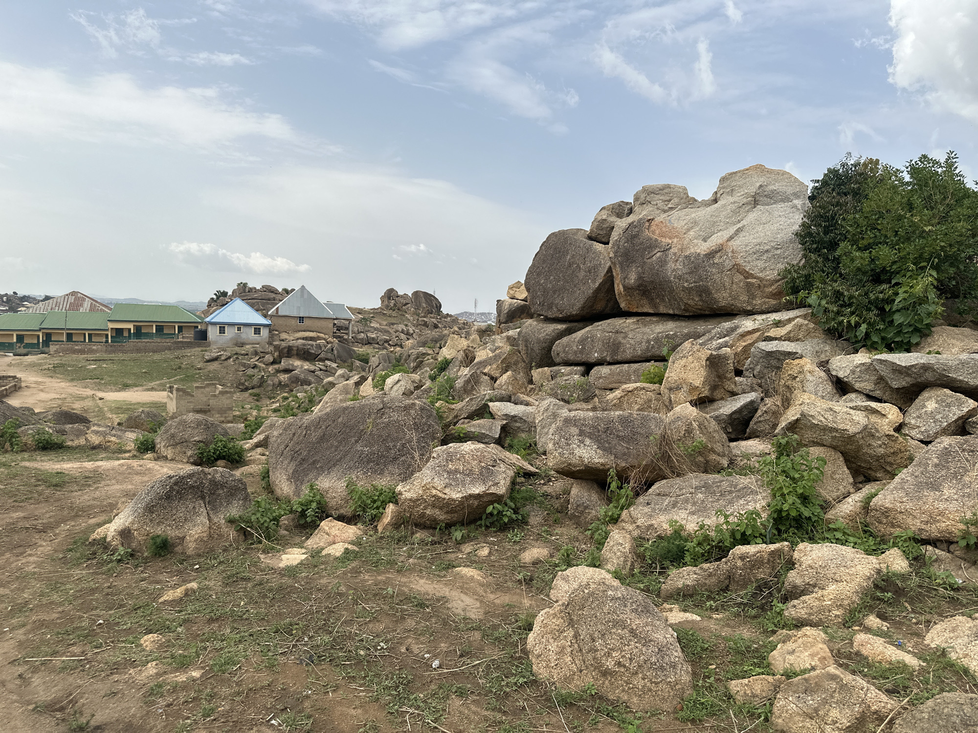 The image shows a rocky landscape with scattered boulders and sparse vegetation in Jos, Nigeria. In the background, a series of low-rise buildings with varying roof colors (green, blue, and grey) can be seen, suggesting a residential or institutional area. The foreground is dominated by a cluster of large, light-colored boulders, some piled atop each other. Small patches of grass and weeds grow between the rocks. The sky is overcast with scattered clouds. No people are visible.