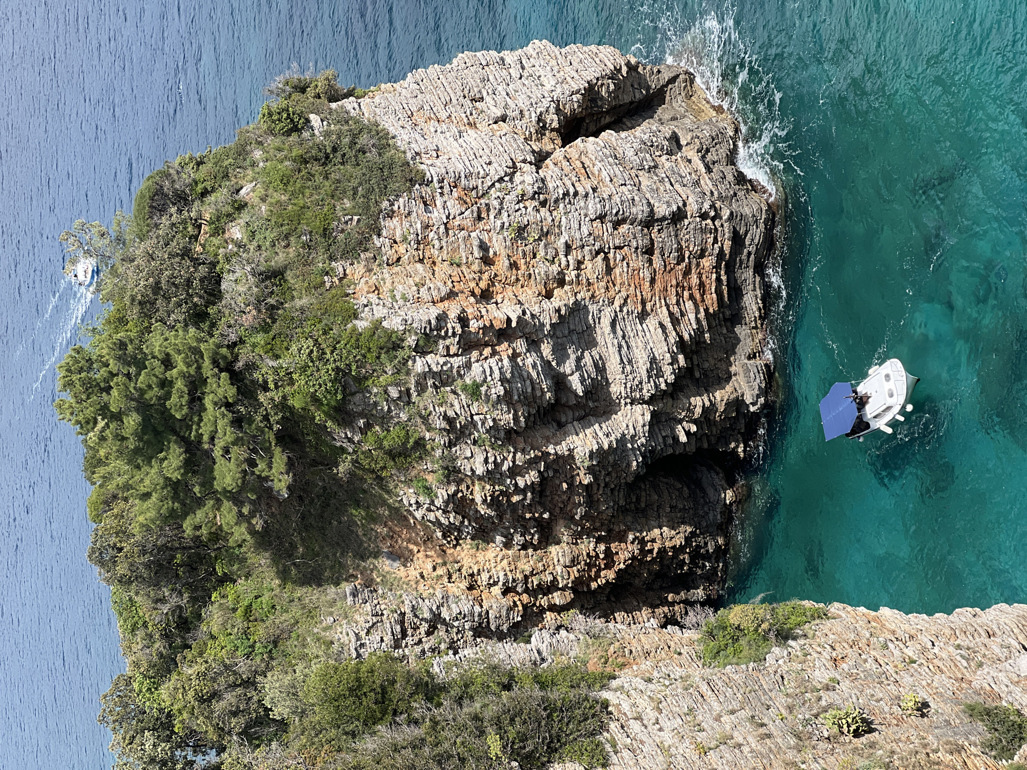 A high-angle shot depicts a rocky coastal landscape in Budva, Montenegro. The scene is dominated by a large, layered rock formation with varied coloration ranging from light gray to brown. Vegetation, including green trees and shrubs, covers parts of the rock. Adjacent to the rock formation is clear turquoise water with a small white boat and a blue awning is positioned. Another boat can be seen further in the distance. The body of water extends to the upper left, displaying a calmer, darker blue surface. The environment is rocky and the weather appears sunny and clear.