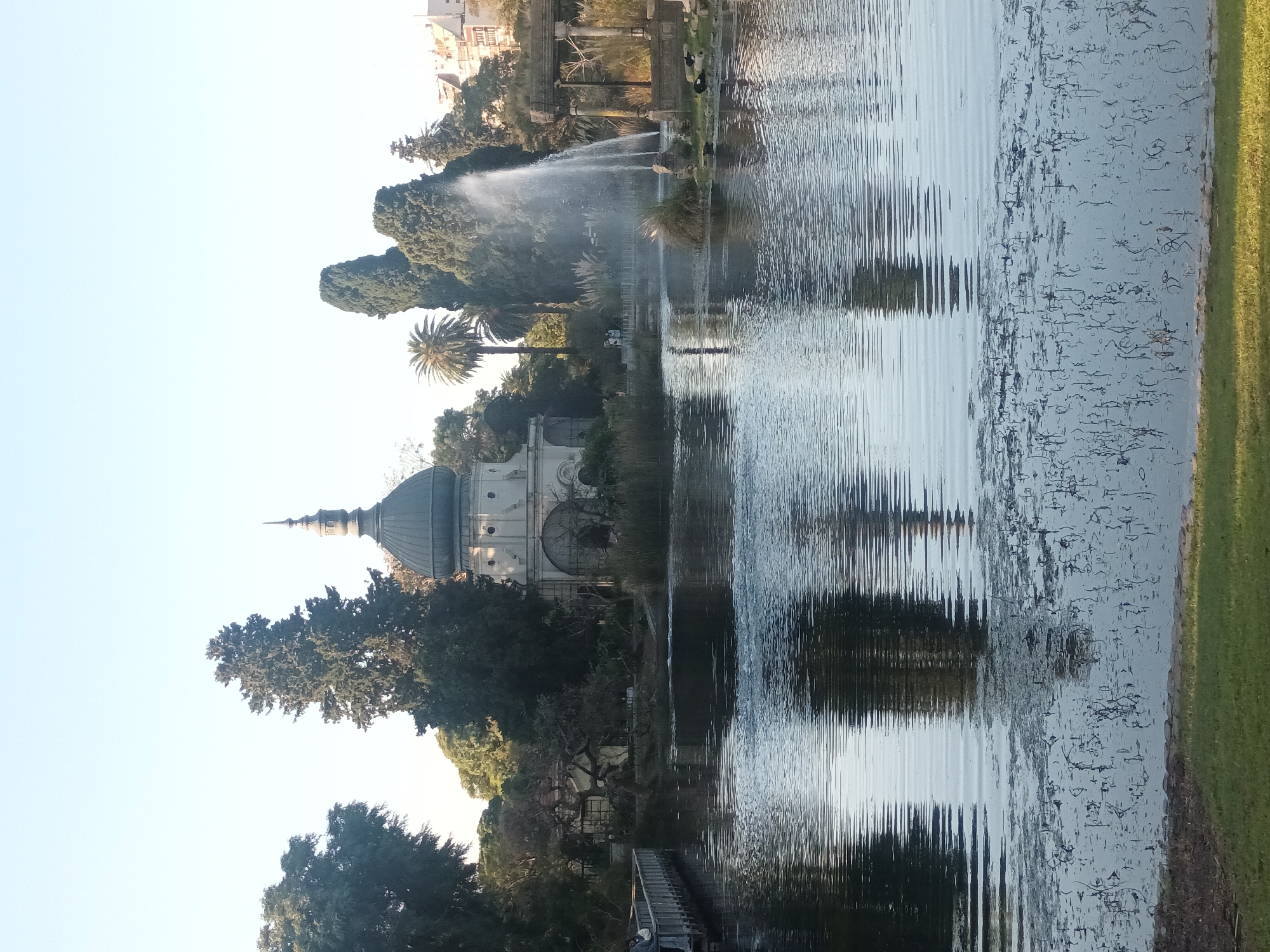 This image shows a view of the Rosedal de Palermo in Buenos Aires, Argentina, with a focus on the lake. The water has visible ripples and reflections of the surrounding architecture and trees. A fountain is visible near the center of the lake. The dome of a building with light-colored exterior and darker dome sits among trees. The sky is a pale blue. A green patch of grass is visible along the bottom right.