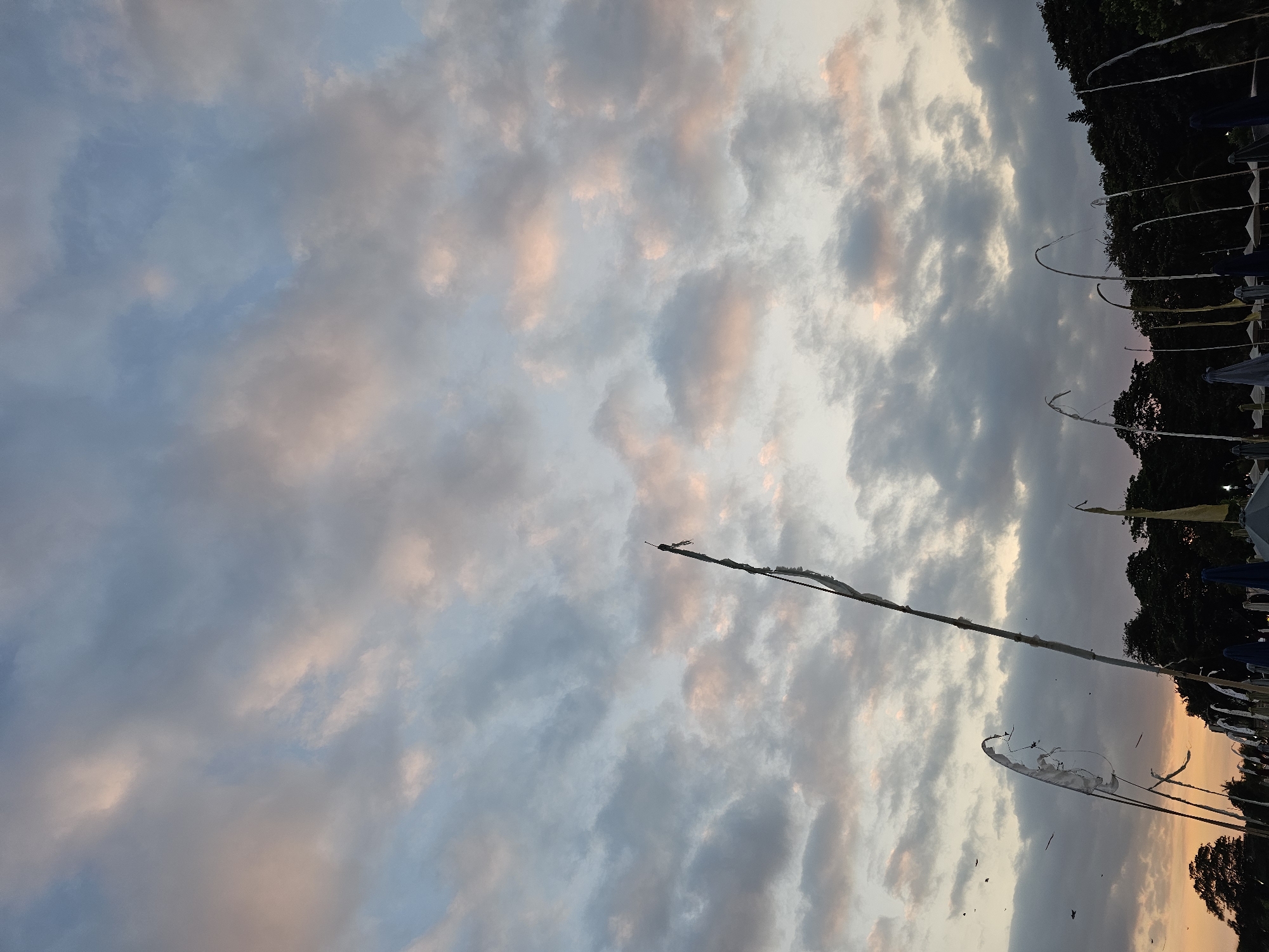 This is a photograph of a cloudy sky over a beach in Denpasar, Indonesia, during sunset. The clouds are a mix of gray and pink hues, indicating the sun is low on the horizon. In the foreground, there are several tall bamboo poles with white tattered flags or streamers attached, and further back, a line of trees and beach umbrellas are visible. There are no people clearly visible in the frame. The overall atmosphere is serene, with the fading light casting a warm glow over the scene. The shot is taken from a low angle, looking up at the sky and the decorative elements.