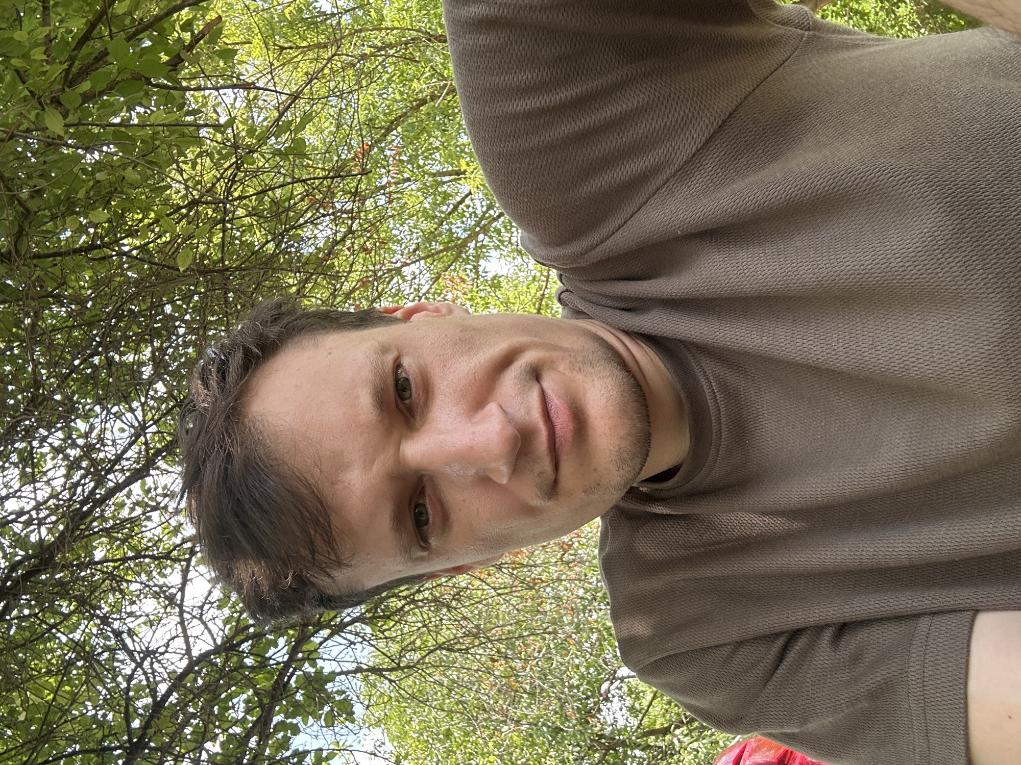 A man is taking a selfie outdoors. He is wearing a brown, short-sleeved, textured shirt. He has short, dark brown hair and light-colored eyes. The background is filled with green foliage and branches, suggesting he is under a tree. The lighting indicates it is daytime. No specific location in the city of Makiv, Ukraine, can be determined from the image.