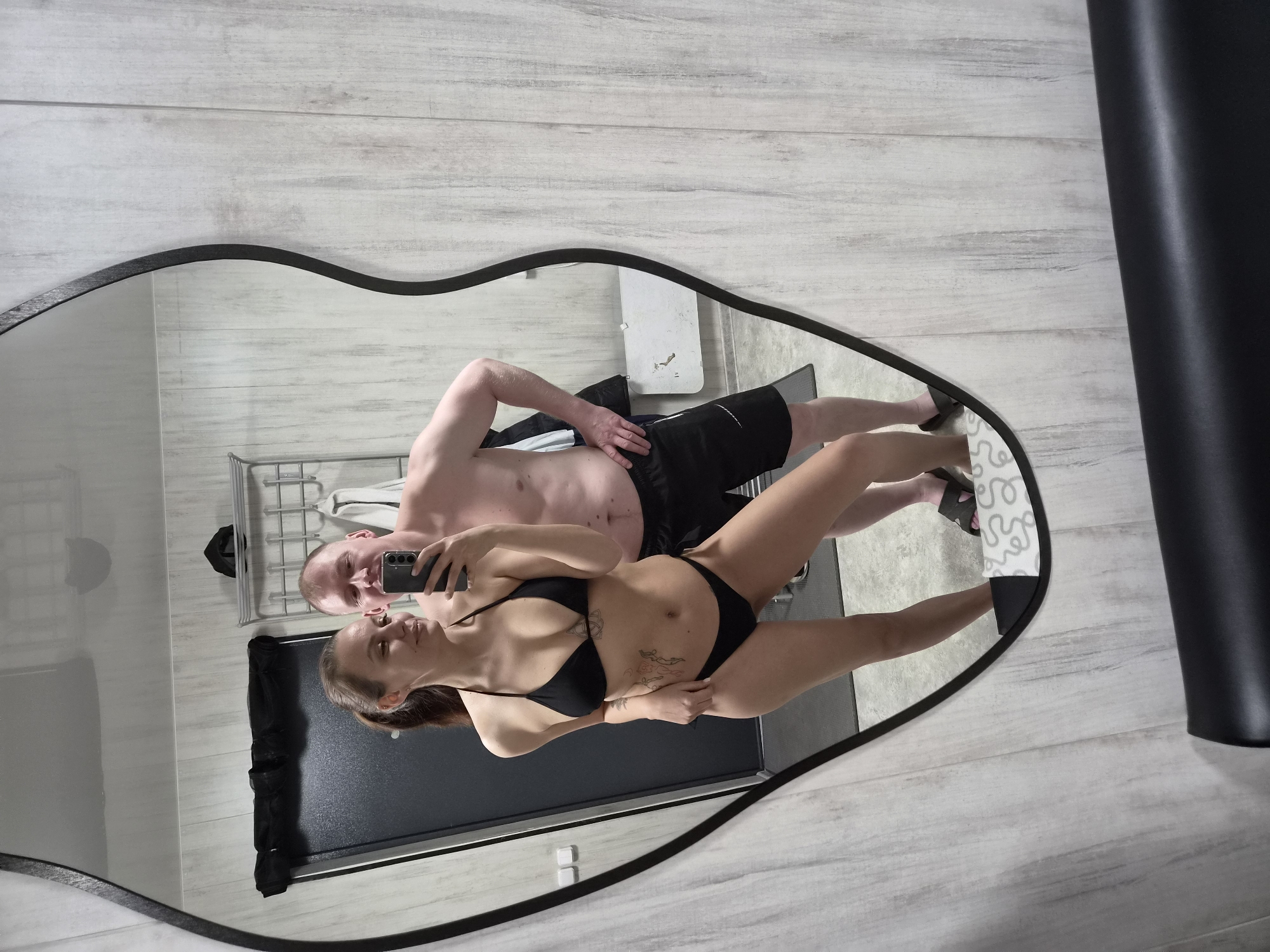 This mirror selfie, taken in Chaikovsky, Russia, features a man and a woman reflected in an irregularly shaped, black-framed mirror. The woman, positioned in the foreground of the reflection, is holding a black smartphone to capture the image. She is wearing a black bikini and has dark hair, likely tied back, with a tattoo visible on her left side/abdomen. She smiles subtly towards the camera.
Behind her, a man is partially visible, also looking into the mirror with a slight smile. He is shirtless and wears black shorts, with one hand casually placed on his hip.
The setting appears to be a bathroom or changing room, characterized by light-colored, wood-look tiled walls and floor. In the background of the reflection, a metal shelving unit is visible behind the man, alongside a dark gray or black surface that could be a shower tray or bench. A white mat with a subtle gray, swirling pattern is under the man's feet. To the far right, outside the mirror's frame, a black object or curtain is partially seen. The atmosphere conveyed is relaxed and personal, capturing a moment between the two individuals. No legible text is visible within the image.