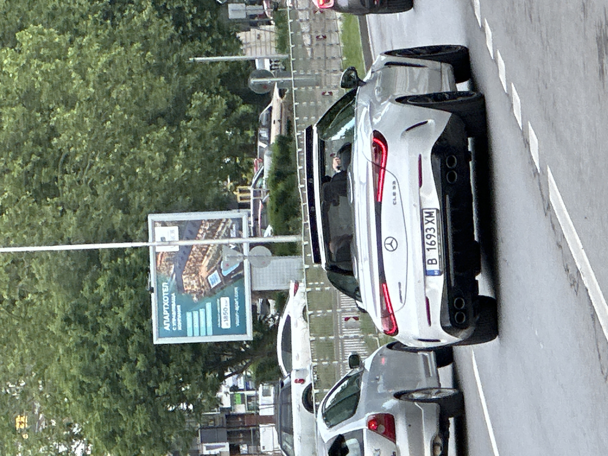 The image shows a white Mercedes-Benz parked on a city street in Varna, Bulgaria. The car's license plate is B 1693 XM. A person is seated in the driver's seat. A billboard advertising "Apart Hotel" is visible in the background. Other cars are parked nearby. The environment includes trees and urban infrastructure like traffic lights and power lines. The street appears paved and wet, with lane markings visible.