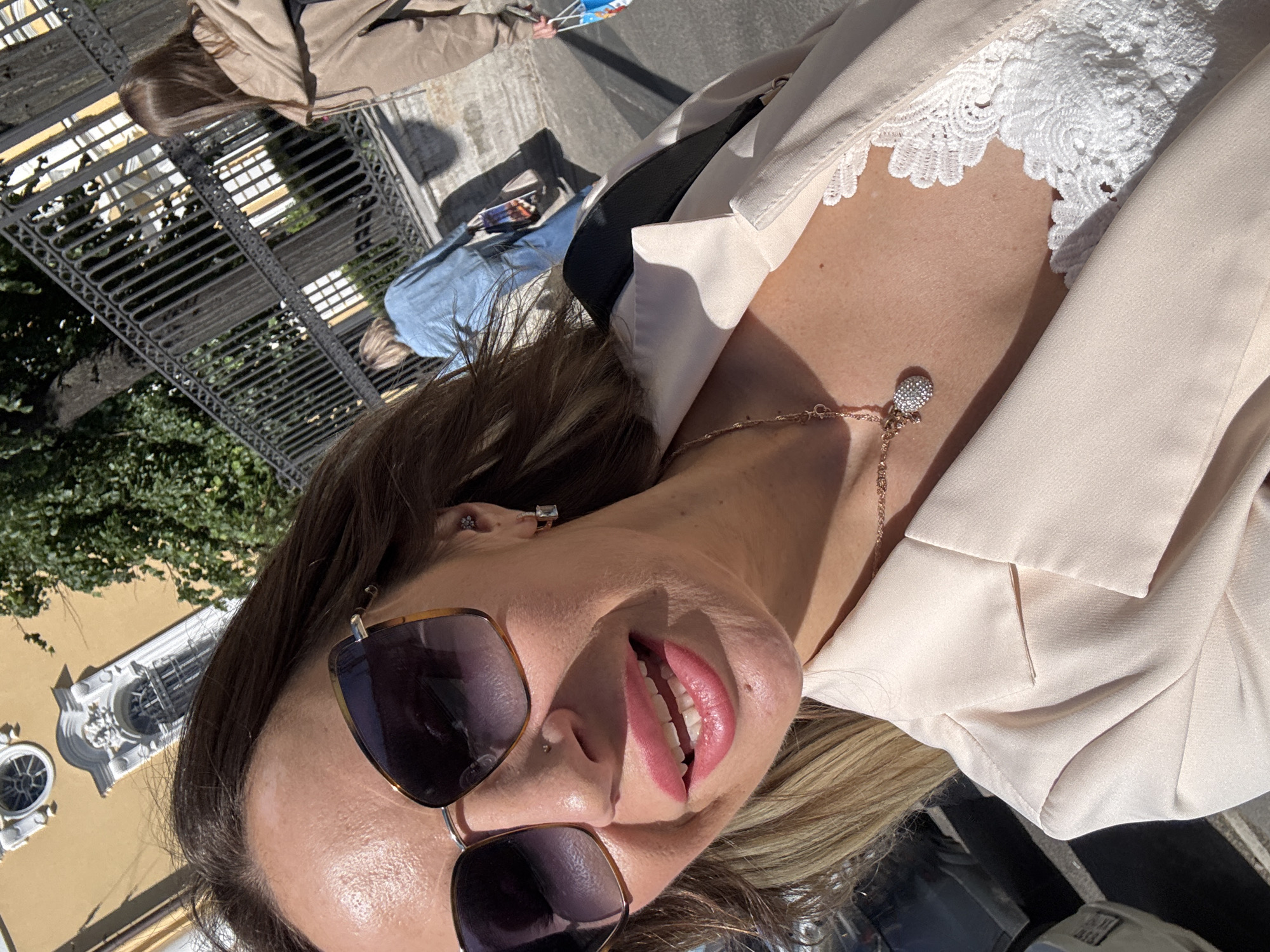 The media file shows a close-up, angled selfie of a woman outdoors on a sunny day. She is wearing a light-colored blazer over a white lace top and a delicate rose-gold necklace with a small, sparkling pendant. Her lips are parted, revealing white teeth, and she is wearing large, dark sunglasses. The background shows a building with ornate architectural details, suggesting a historic European city. Trees and a metal fence are also visible. In the further background, other people are seen, one of whom is wearing a blue denim jacket. The lighting indicates it is daytime, likely a bright, sunny afternoon. The woman's facial expression could be interpreted as playful or inviting. There is no visible text in the image. The context provided indicates the location is Saint Petersburg, Russia.