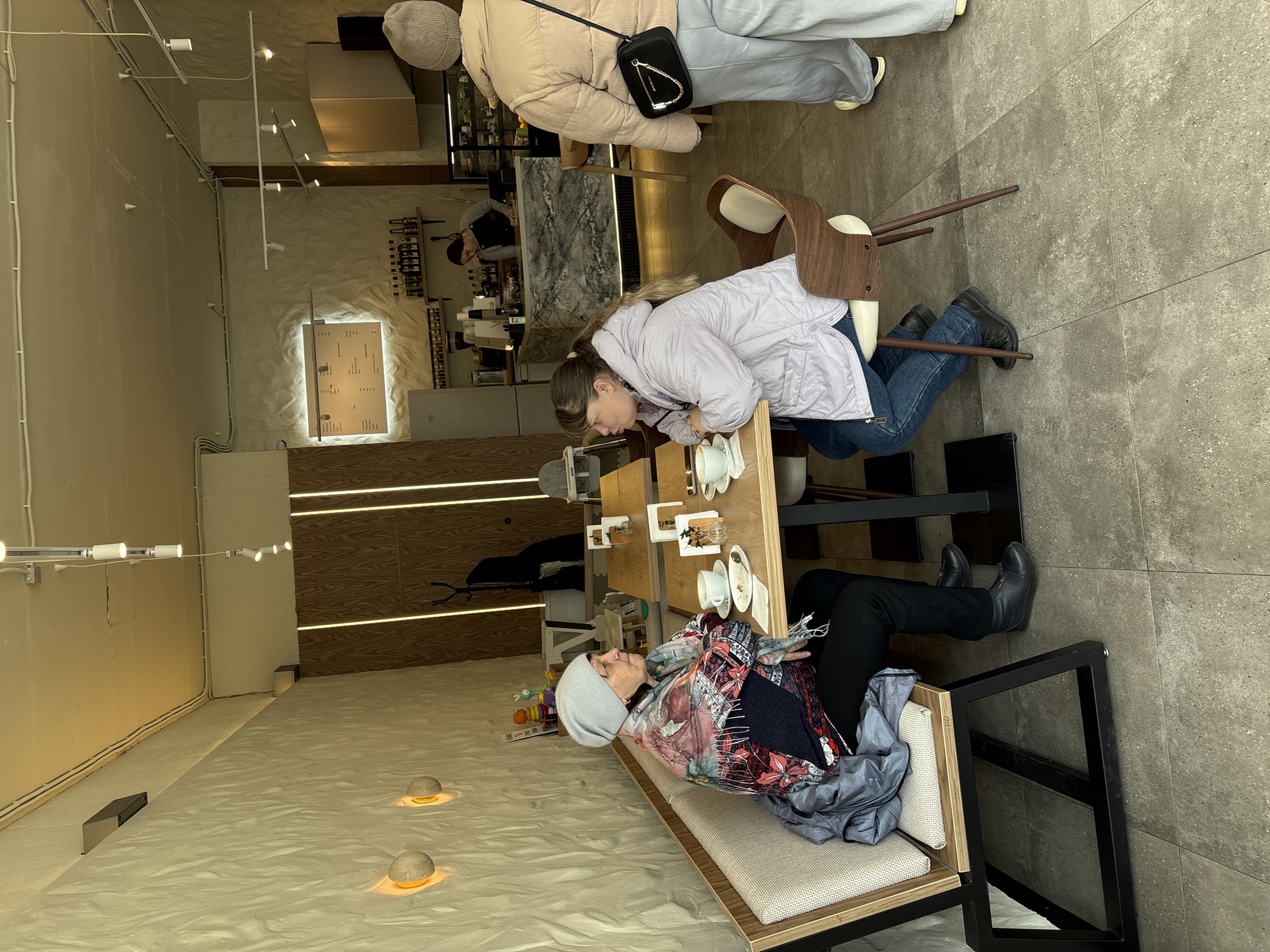 The image depicts the interior of a cafe. Two women are seated at a table, seemingly engaged in conversation, leaning towards each other with teacups visible on the table. One woman wears a light purple jacket and jeans, while the other, seated on a cushioned bench, wears a patterned top with a gray hat. Another person stands near a counter further back in the cafe. The cafe's decor includes a patterned wall, wooden accents, and spherical lights on the floor. The floor appears to be light in color, perhaps textured. The location is Ulyanovsk, Russia.