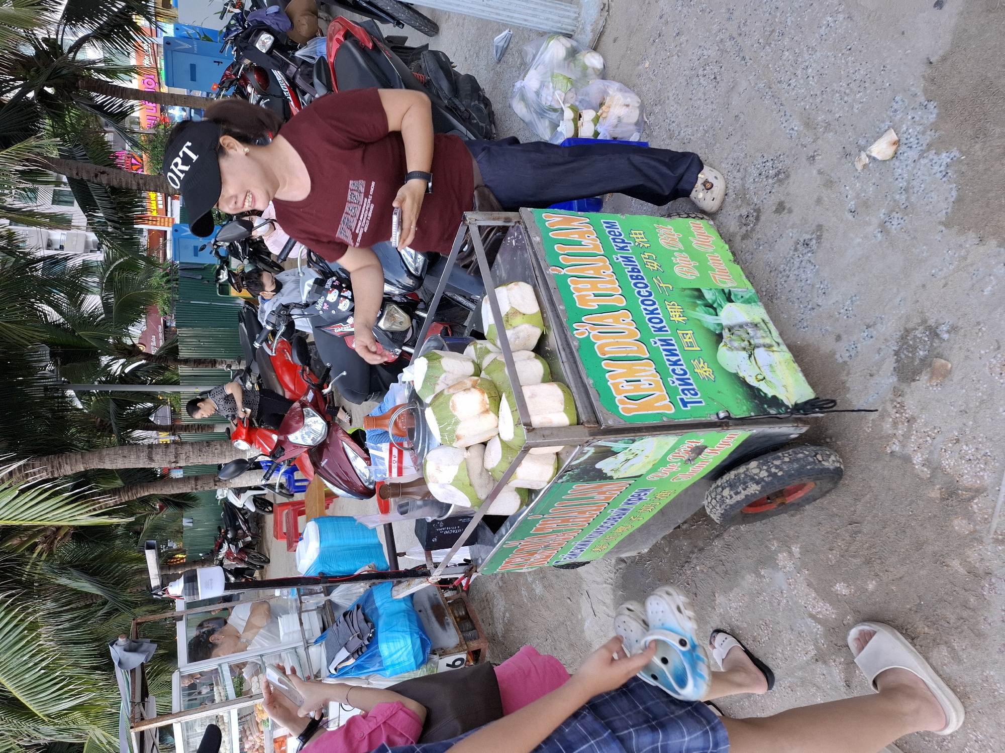 In Bac Nha Trang Ward, Vietnam, a woman stands behind a coconut cart, smiling as she holds a mobile phone. The cart is filled with halved coconuts, and a sign in green displays Vietnamese and Russian text, indicating "Thai coconut cream." To her left, a person wearing a pink shirt and blue shorts is partially visible, their hand reaching towards the cart. In the background, a row of parked motorcycles and palm trees line the sandy street. The scene appears to be a bustling street market or a roadside vendor area.