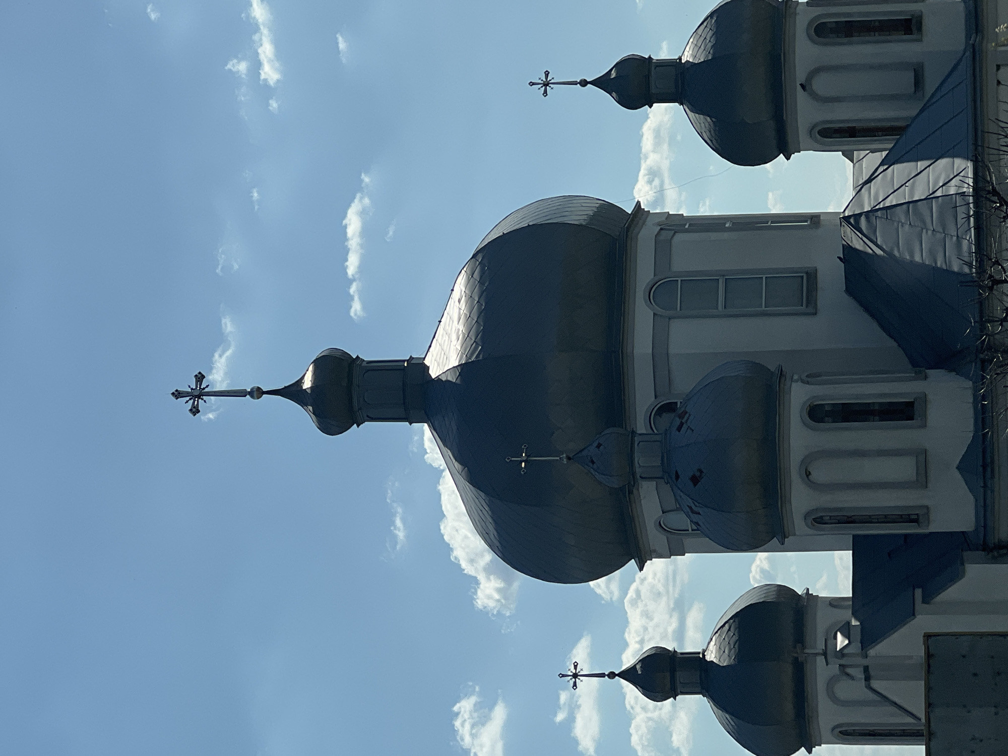 The image features a low-angle view of a church with multiple domes under a partly cloudy, bright blue sky. The domes are dark and appear metallic, topped with crosses. The church walls are light-colored, possibly white or light gray, with architectural details like windows. The location of the church is Bila, Ukraine. The image composition is such that the church is viewed from below, emphasizing its height and grandeur against the sky.