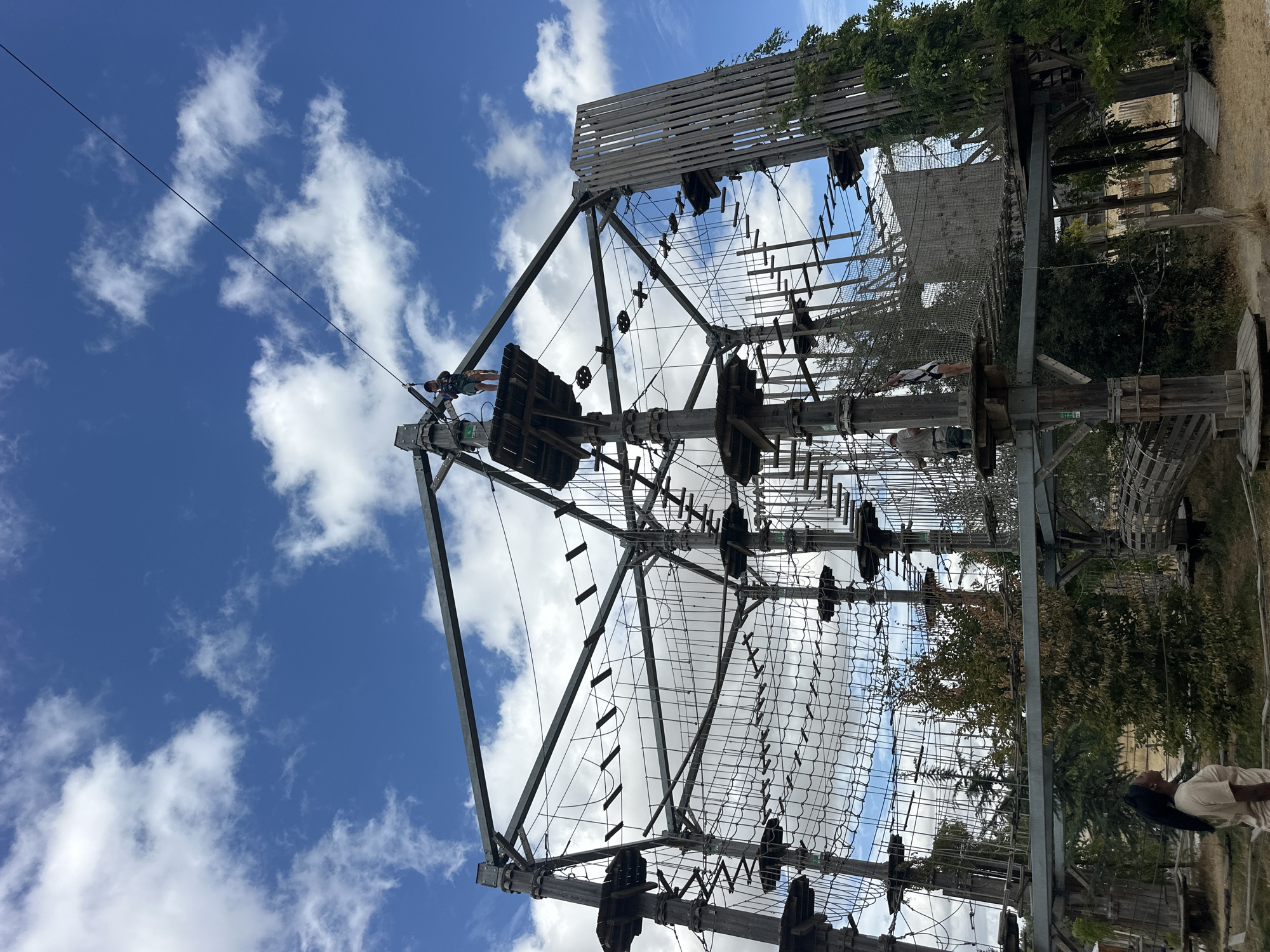 The image captures an expansive adventure park set against a bright blue sky with scattered white clouds. The park features a large, multi-level obstacle course constructed from wood and metal, with various suspended bridges, ropes, and platforms. Several individuals are visible navigating the course. In the foreground, a woman with dark hair and a light-colored t-shirt stands looking upwards and to her left. Further into the course, a young boy in a green and black t-shirt and shorts stands on a platform, attached to a safety line. Another individual, wearing a white shirt and shorts, is on a net bridge further into the course. The surrounding environment includes trees and dry ground, suggesting an outdoor recreational setting. The location is identified as Jablines, France.