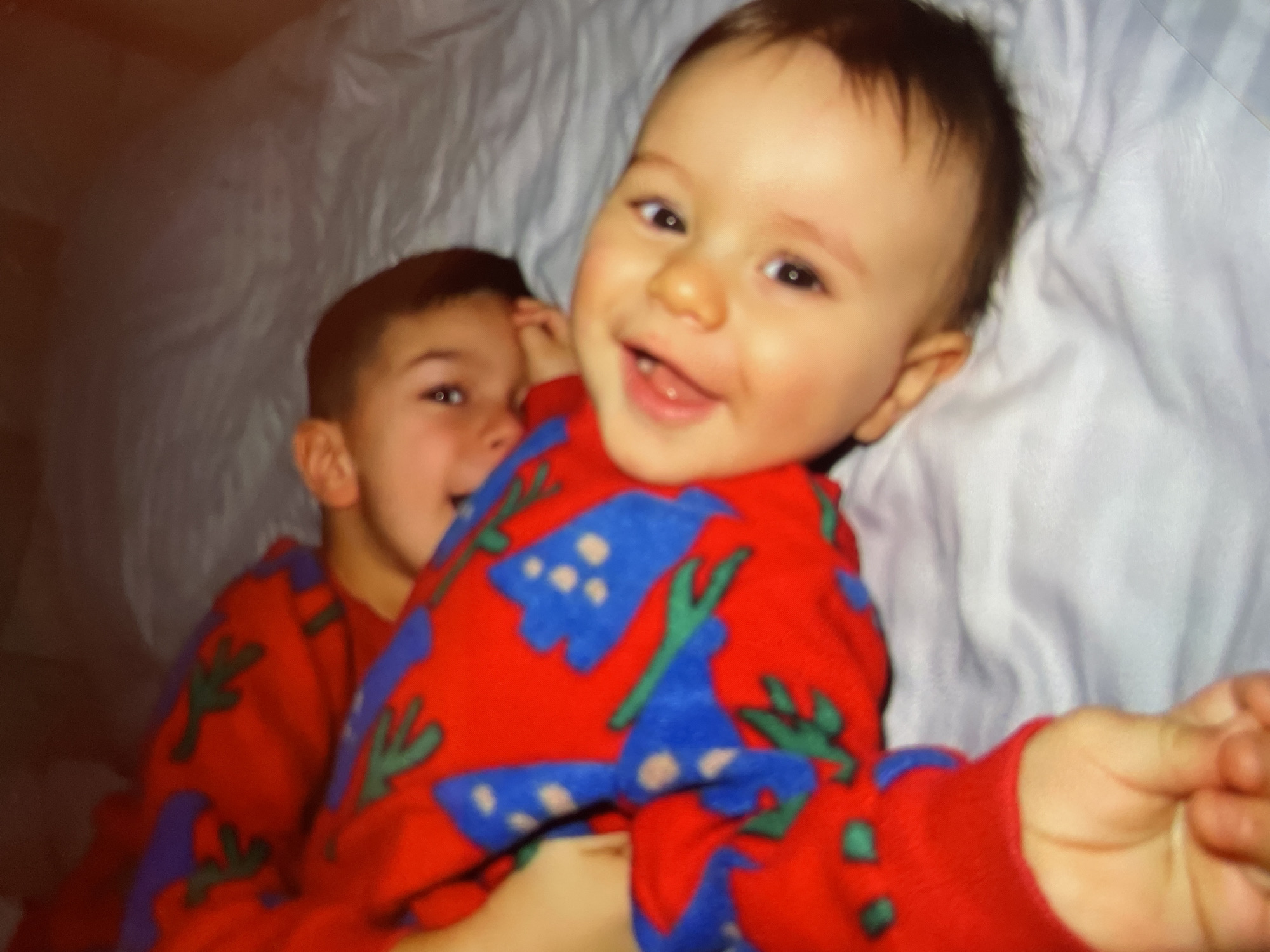 Two young children are pictured. The child in the foreground is smiling, with an open mouth showing teeth. Both children are wearing red pajamas with a blue and green pattern. They are lying on a white blanket. The location is Harmanli, Bulgaria.