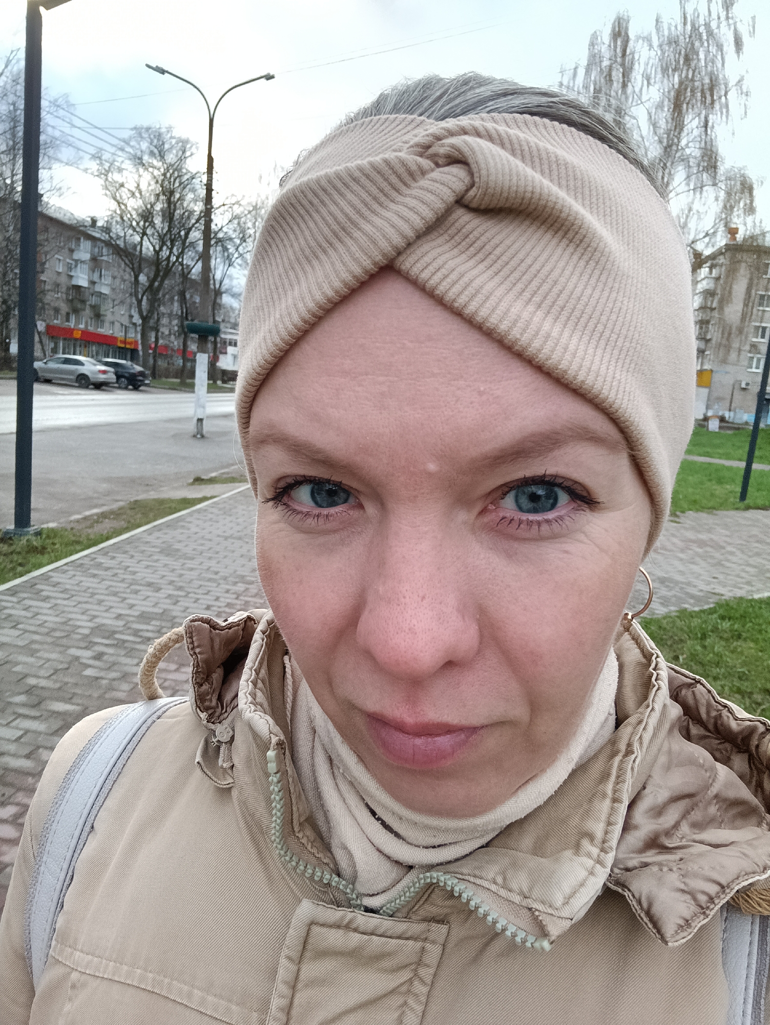 A close-up outdoor selfie or portrait features a woman looking directly at the camera. She has blue eyes and fair skin, with a neutral to slightly serious expression. Her hair, which appears gray or silver, is mostly covered by a beige, ribbed, knotted headband. She is wearing a light beige turtleneck and a tan or beige utility-style jacket with a prominent zipper that has a light green pull tab. A small gold hoop earring is visible on her right ear. A light-colored strap, possibly from a backpack or bag, is visible over her left shoulder.
The background depicts an urban street scene in Chaikovsky, Russia. It is an overcast day, indicated by the muted light and bare trees, suggesting late autumn, winter, or early spring weather. The woman's warm clothing supports this. In the midground, there are multi-story residential buildings, characteristic of Soviet-era architecture, and a street with parked cars. A tall street light with two lamps extends into the cloudy sky. A paved sidewalk made of grey bricks and patches of green grass are visible in the foreground. No discernable text is visible in the image.