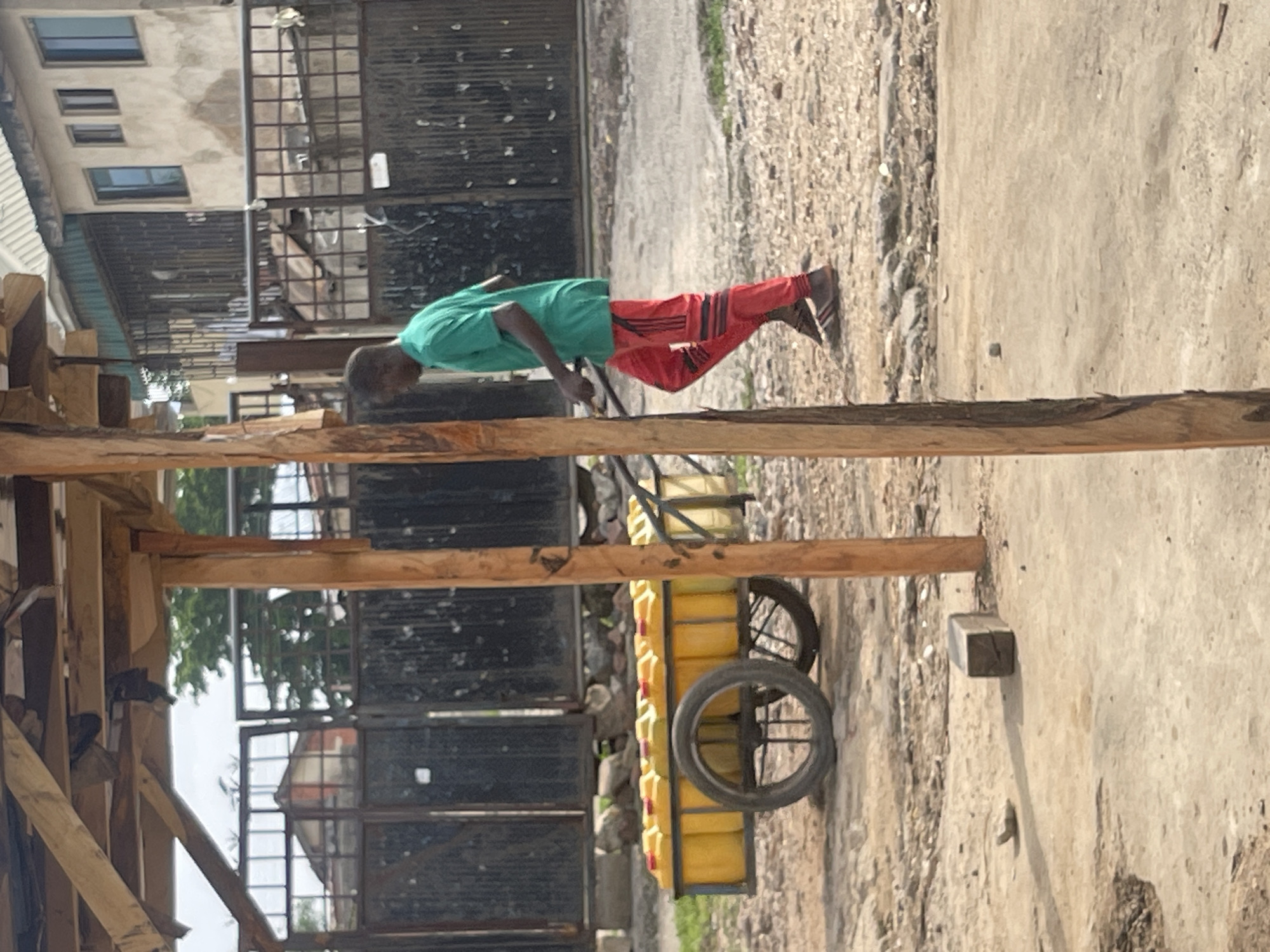An African man wearing a green t-shirt and red pants is pushing a cart filled with yellow plastic containers on a dirt road. The cart has two wheels, one of which is prominently visible in the foreground. The man is walking away from the camera, towards the right of the image. Behind the man and the cart, there is a dark metal fence with a building visible in the distance. The environment appears to be an urban or semi-urban area, possibly a market or a roadside stall. The man is the sole human subject in the image. The lighting suggests it is daytime, with some overcast conditions.